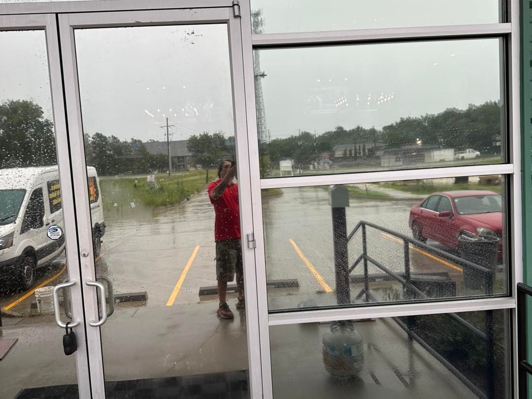 A person in a red shirt stands outside a glass door, observing a parking lot flooded with deep water during heavy rain.