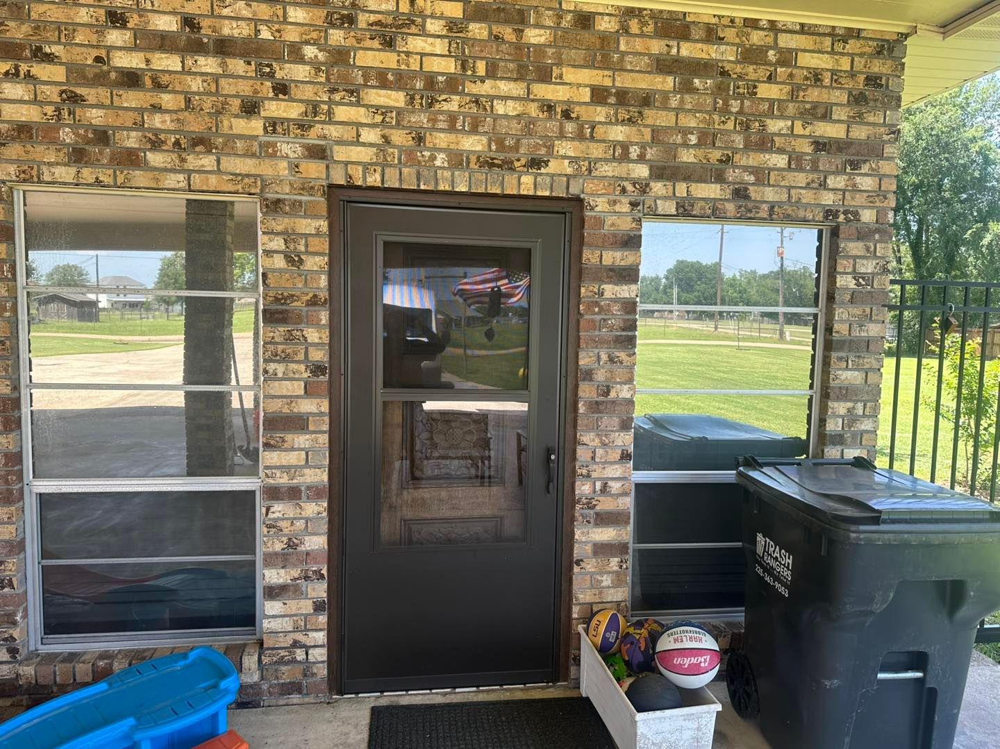 A brick house entryway with a dark storm door, two tall side windows, a blue toy, and a trash bin filled with sports balls.