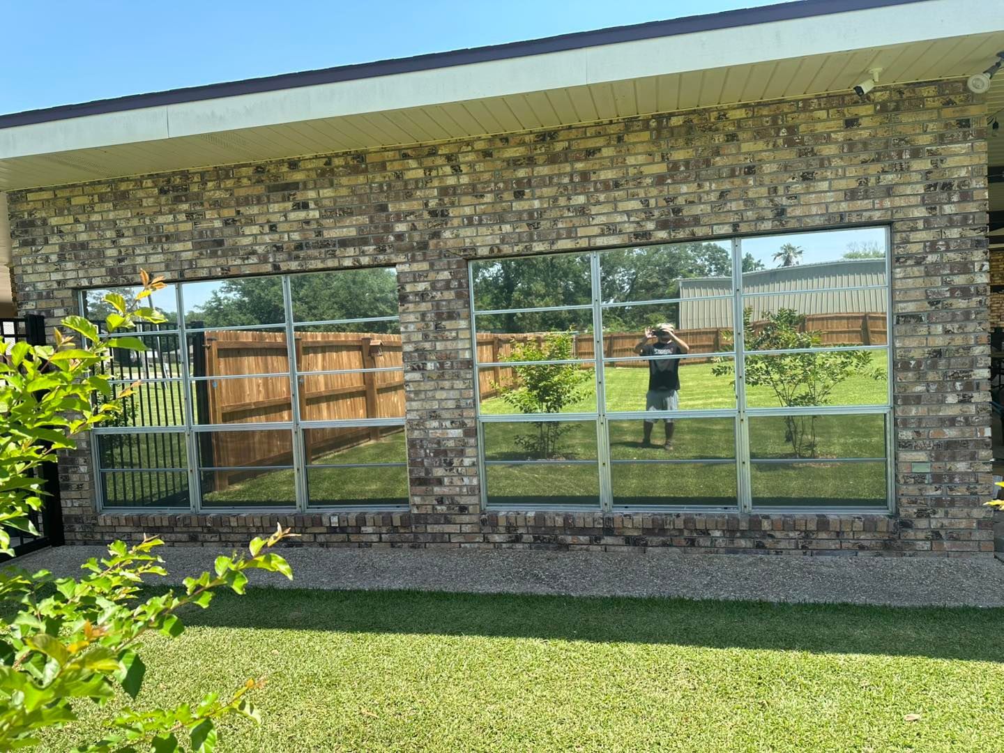 A brick exterior wall features two large glass windows reflecting a lawn, a fence, and a person taking a photo.