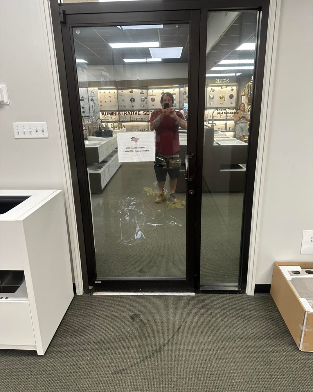 Glass door reflecting a person in a store with shelving units and a sign on the glass.