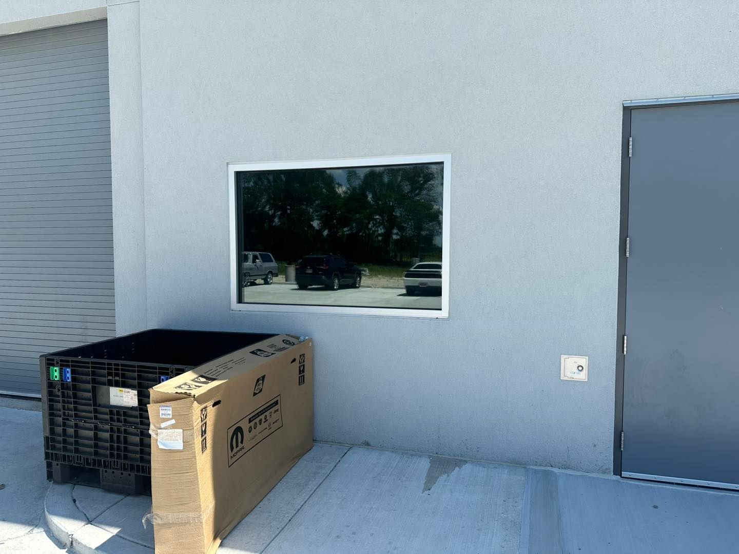 A light gray industrial building wall with a rectangular window, a closed metal door, and a black storage bin near a box.