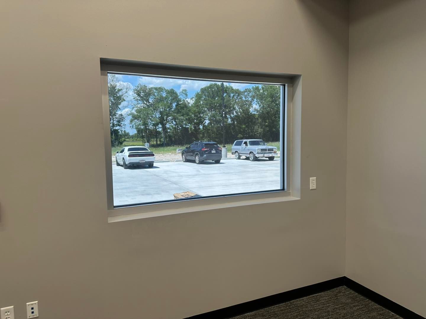 A view from inside a beige room through a rectangular window showing a parking lot with three cars and trees beyond.