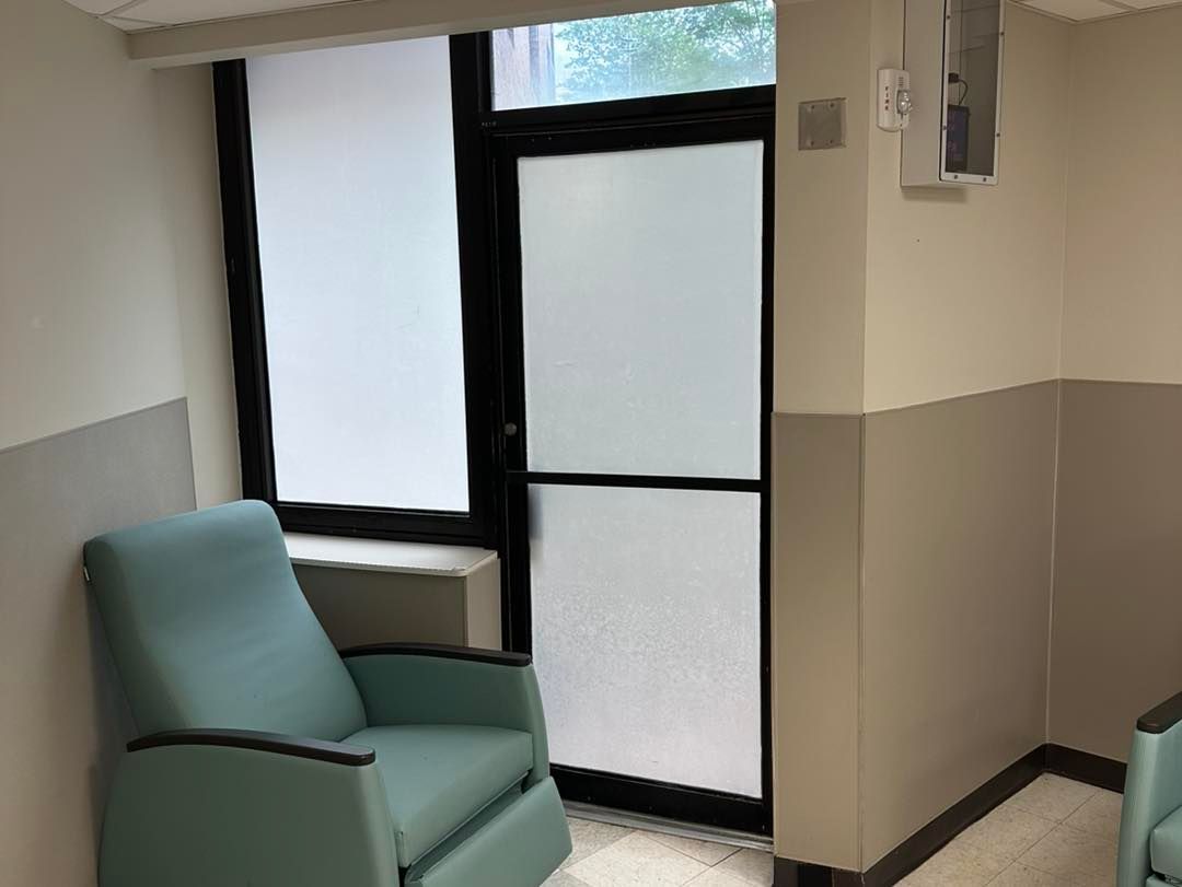 A waiting room with teal armchairs, a frosted glass door, and a window with white blinds against beige painted walls.