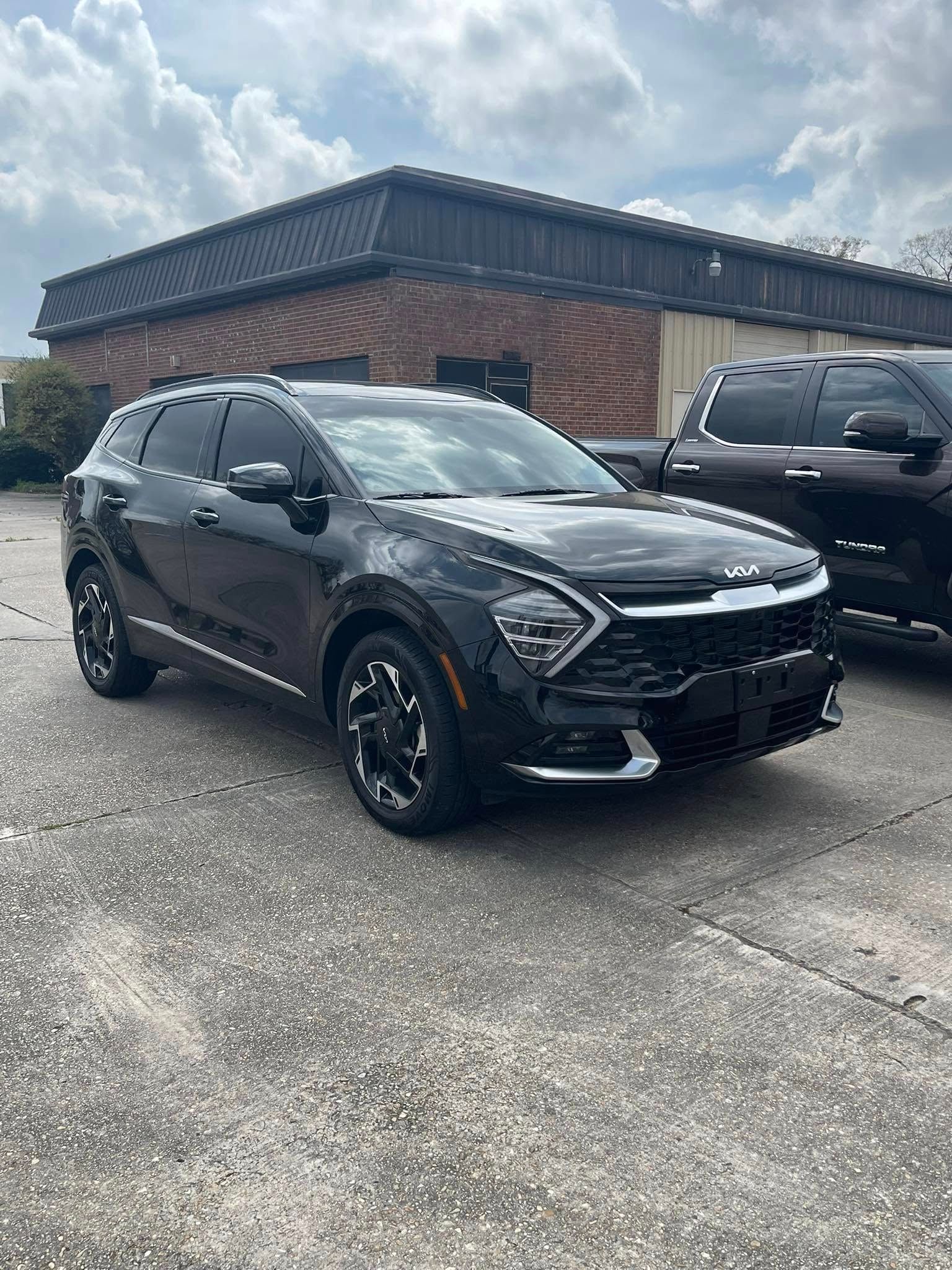 A black Kia Sportage SUV parked on an asphalt lot in front of a brick building under a cloudy sky.