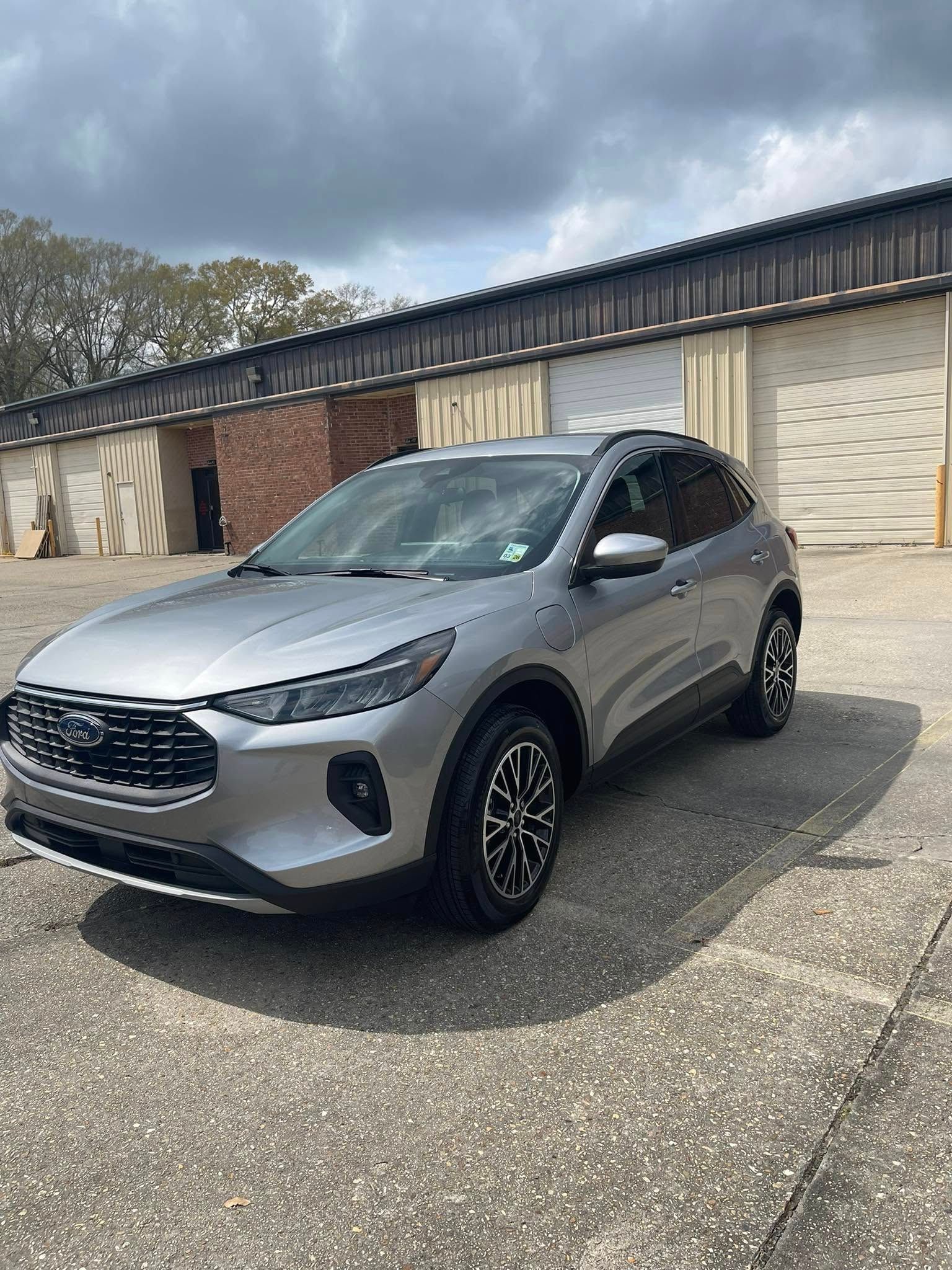 A silver Ford Escape SUV parked on a paved lot in front of a building with multiple garage doors under a cloudy sky.