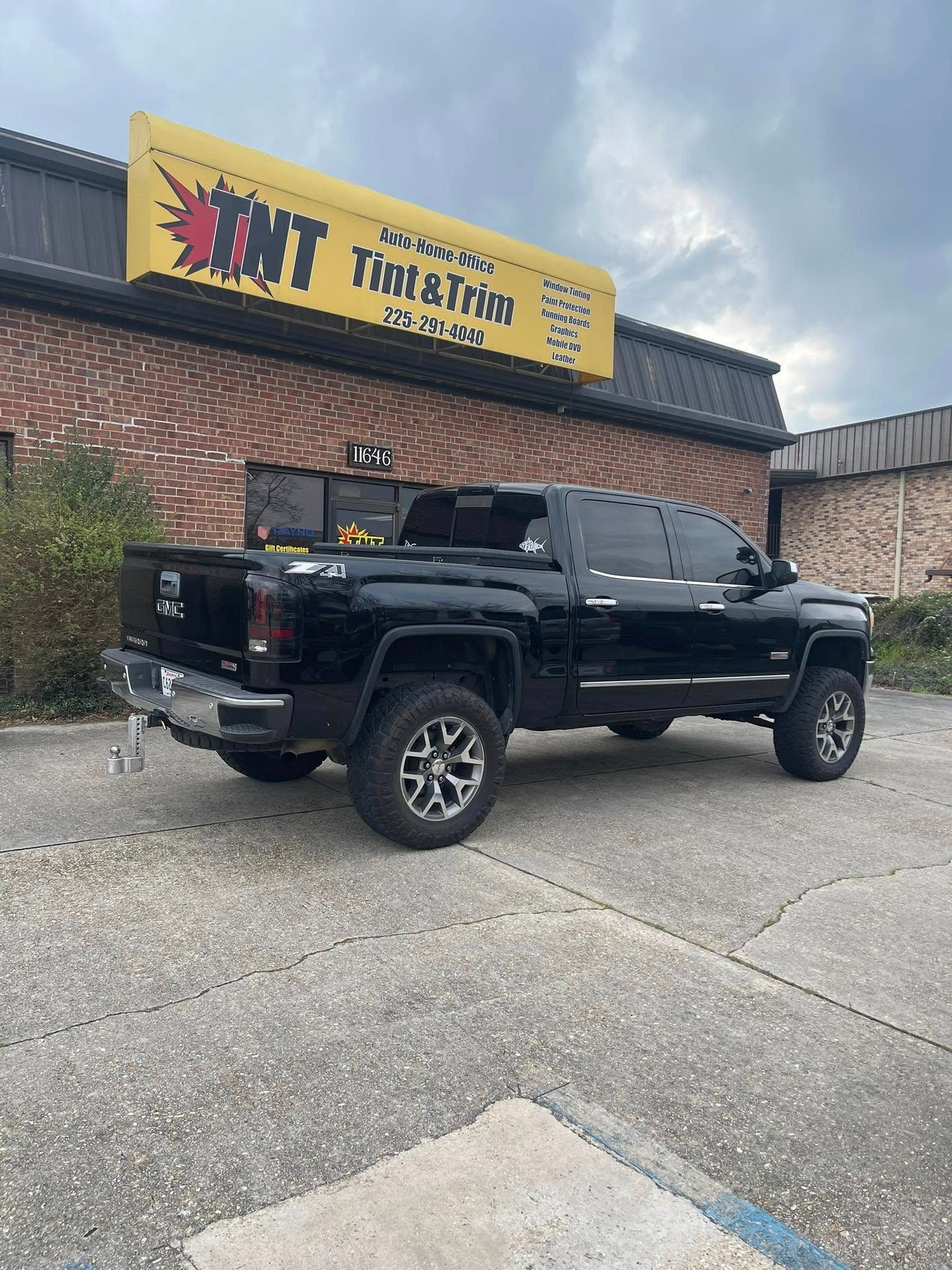 A black lifted pickup truck parked in front of a brick building with a yellow TNT Tint & Trim sign.
