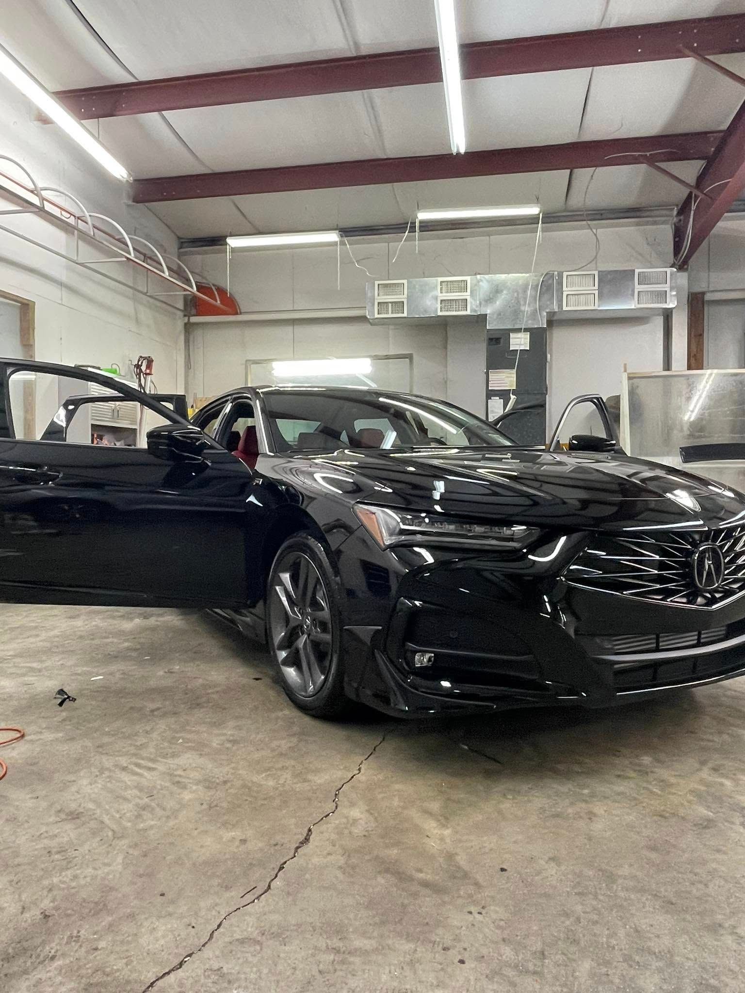 A black Acura sedan with the driver-side door open, parked inside a garage or auto body shop.