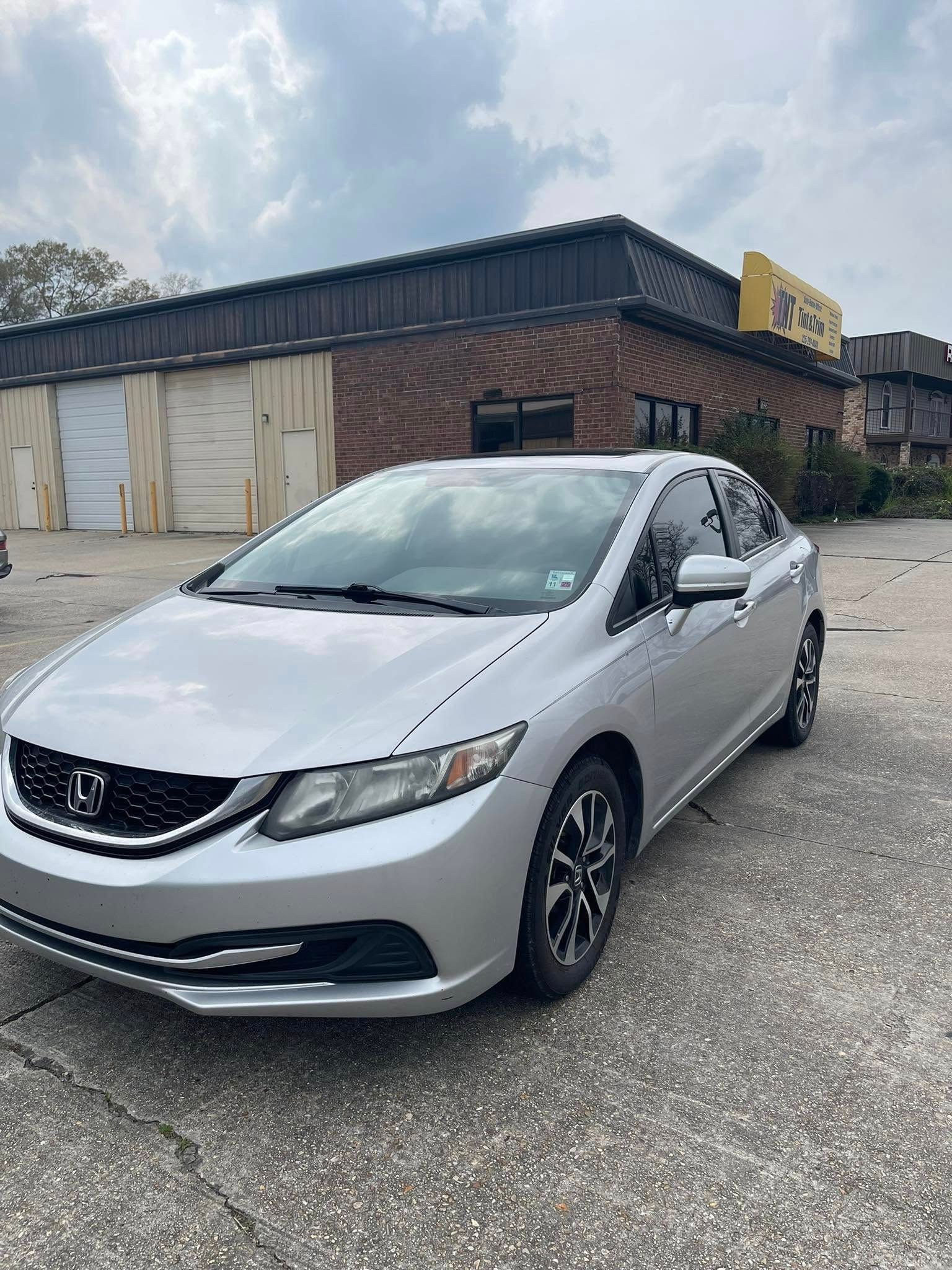 A silver Honda Civic sedan parked in a gravel lot in front of a brick commercial building on a cloudy day.