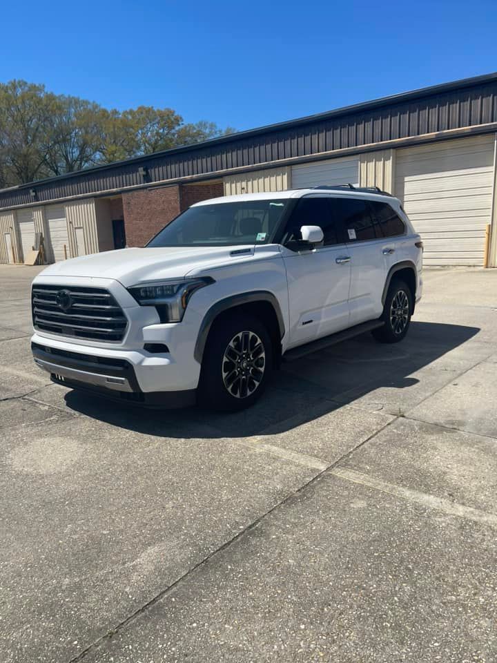 A white Toyota Sequoia SUV parked on a concrete lot in front of a row of storage units under a clear blue sky.