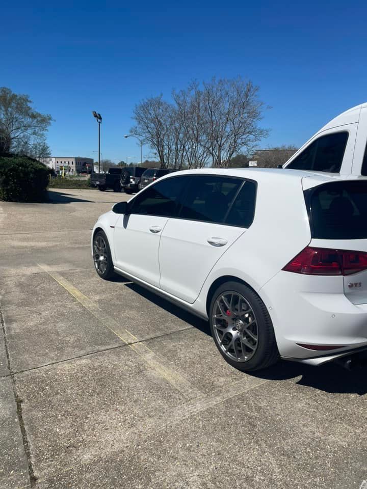 A white Volkswagen Golf GTI parked in a sunny, paved parking lot under a clear blue sky.