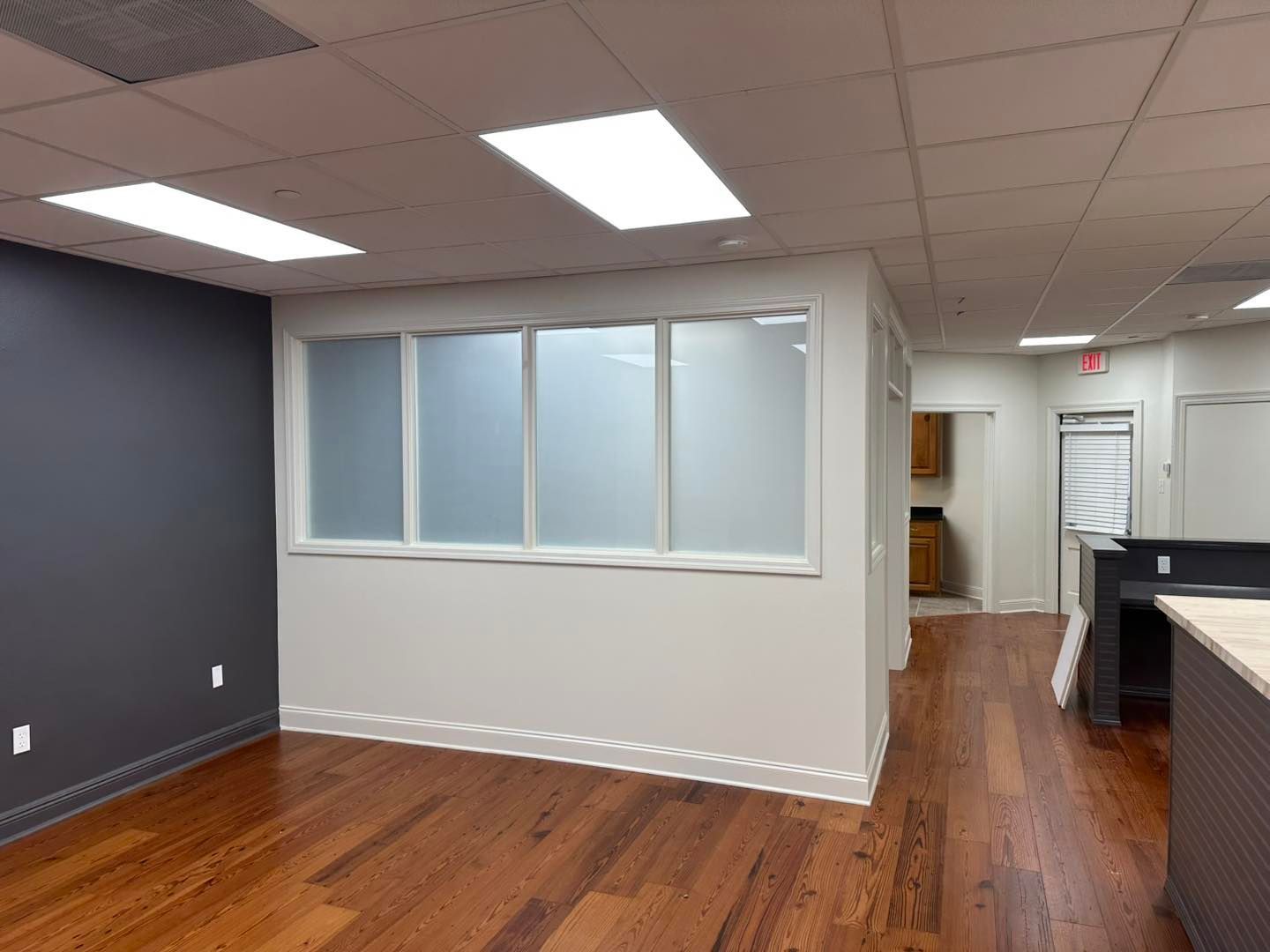An office interior with dark gray and white walls, wood flooring, recessed lighting, and a frosted glass partition.