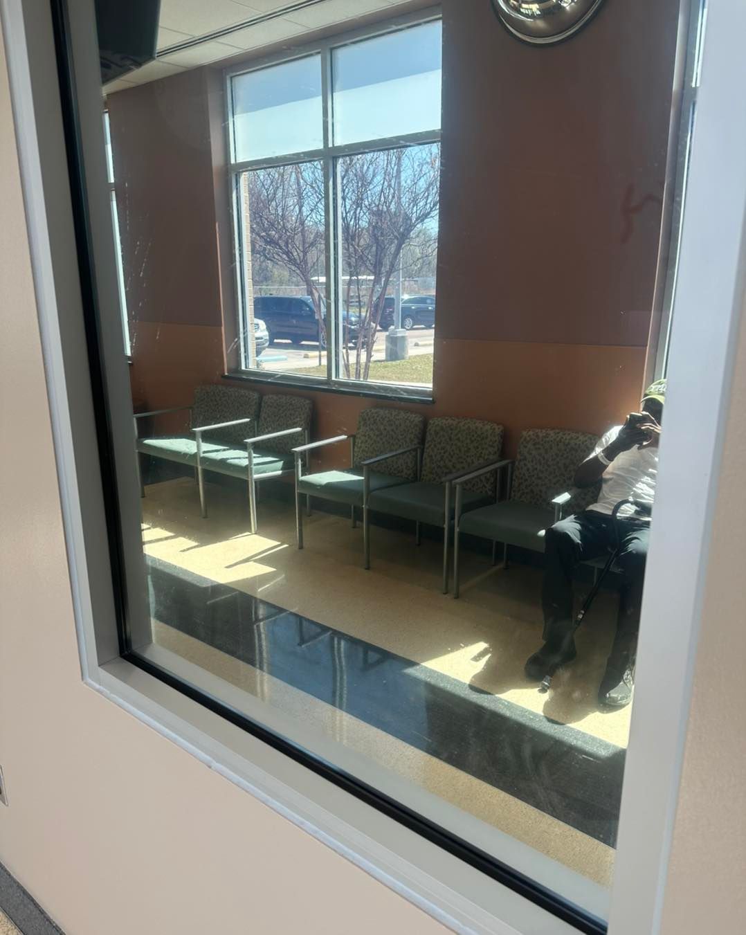 A view through a window into a waiting room with rows of gray chairs and a person sitting on the far right.