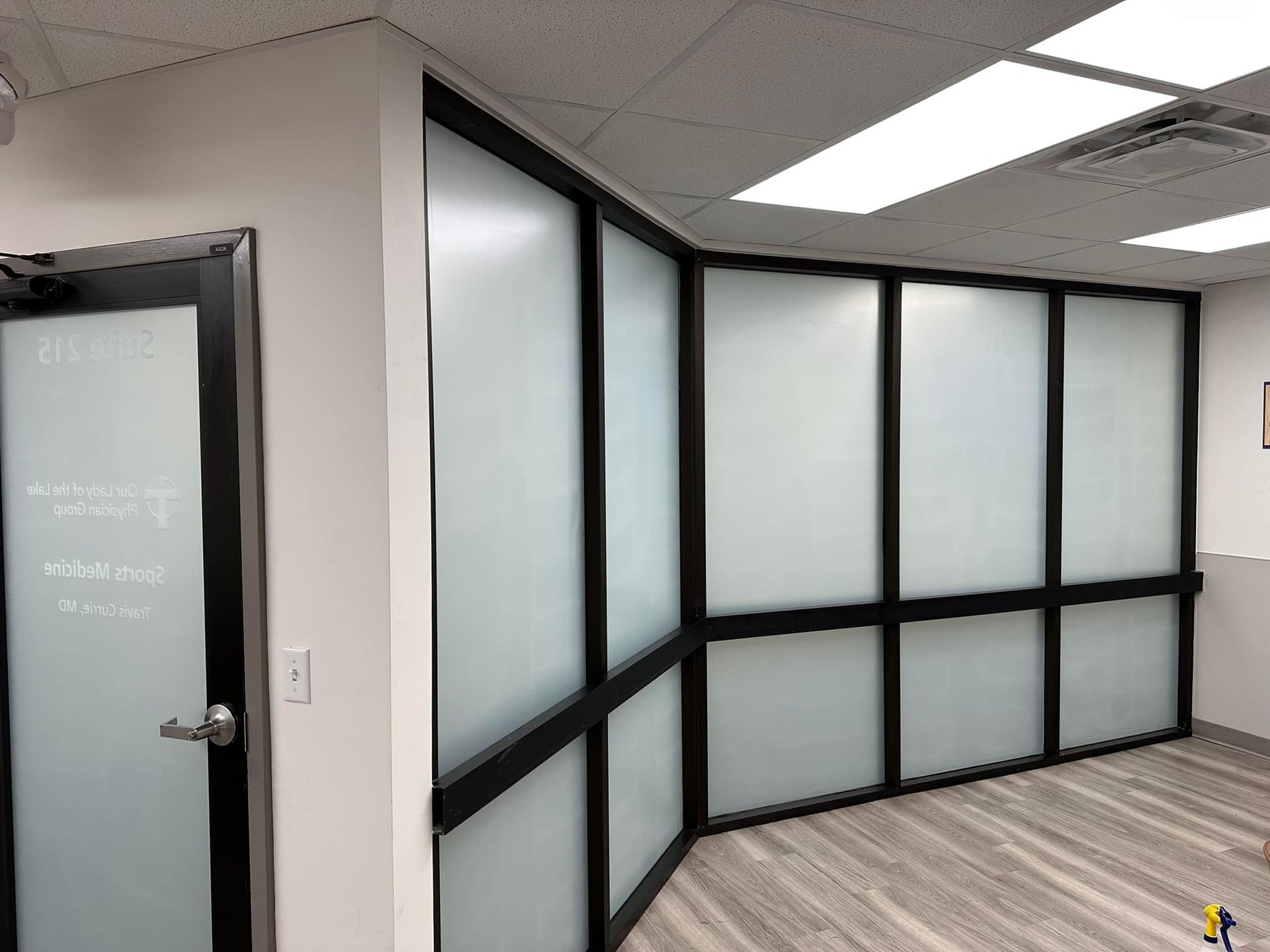 Office interior featuring a glass door and walls with black frames and frosted panels, set on wood-look flooring.
