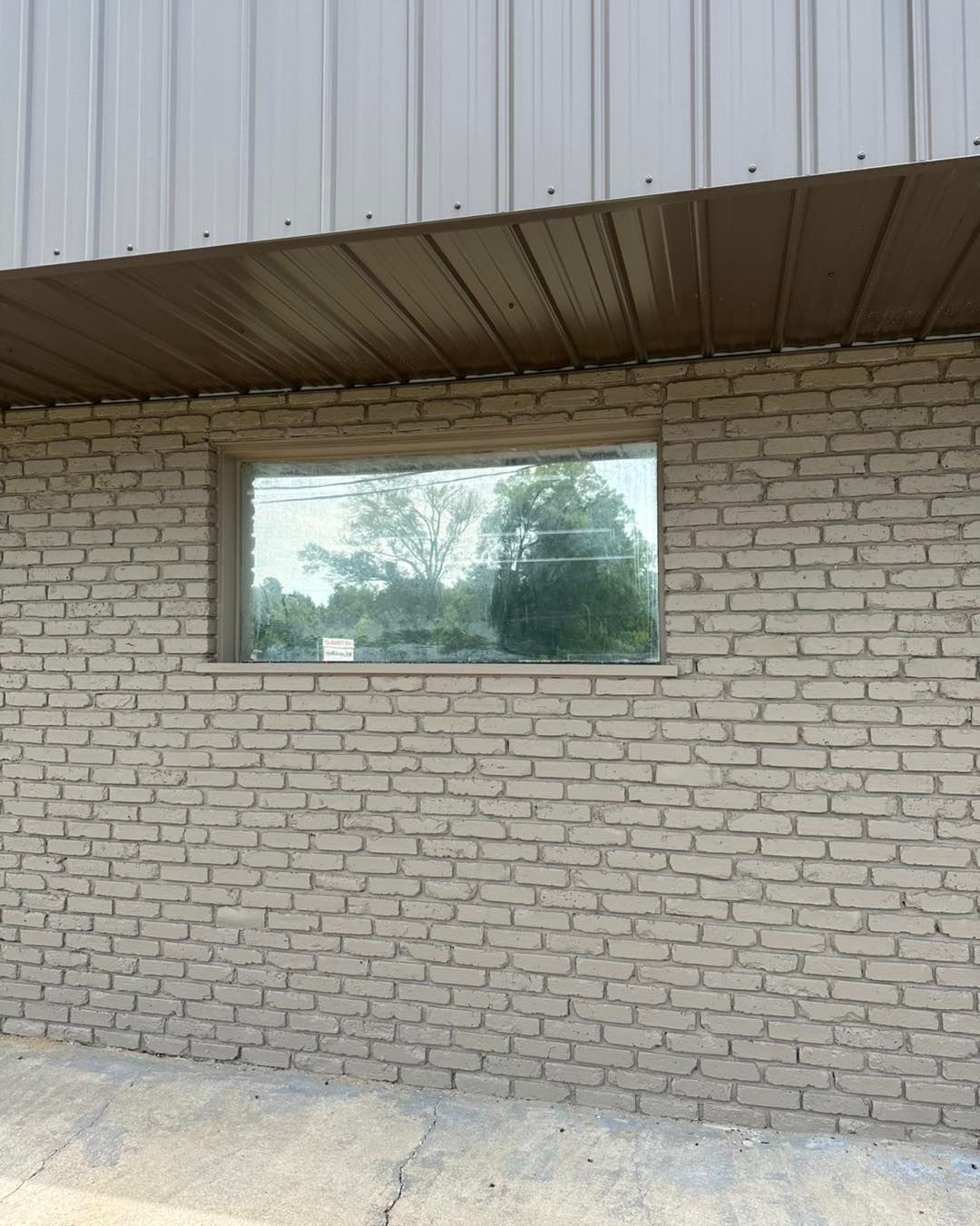 A rectangular window set into a beige brick wall below a metal roof overhang, reflecting trees and a bright sky.