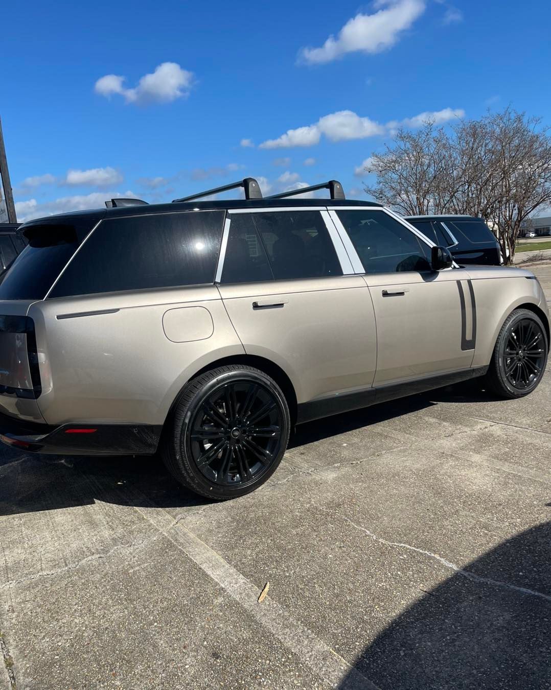 A tan Range Rover with a black roof, roof rails, and black wheels parked on a paved lot under a blue sky.
