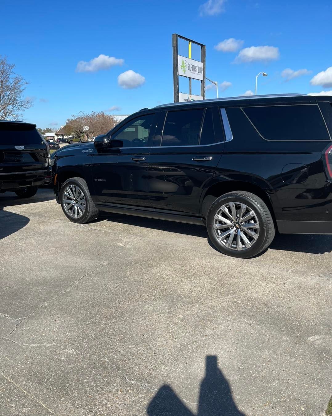 A black SUV parked on a paved lot under a clear blue sky, with a sign and another vehicle partially visible.