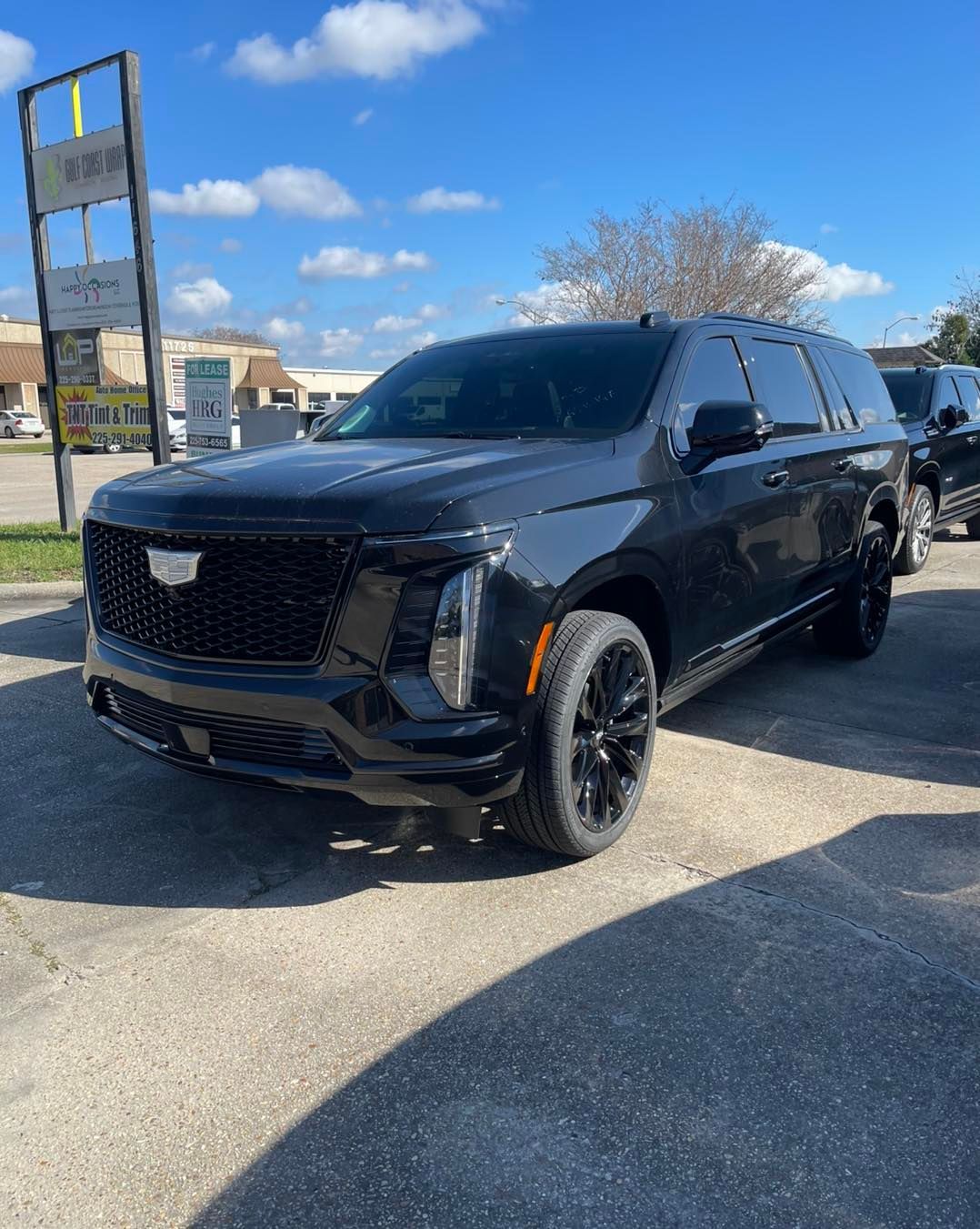 Black Cadillac Escalade parked on a paved lot under a blue, partly cloudy sky.