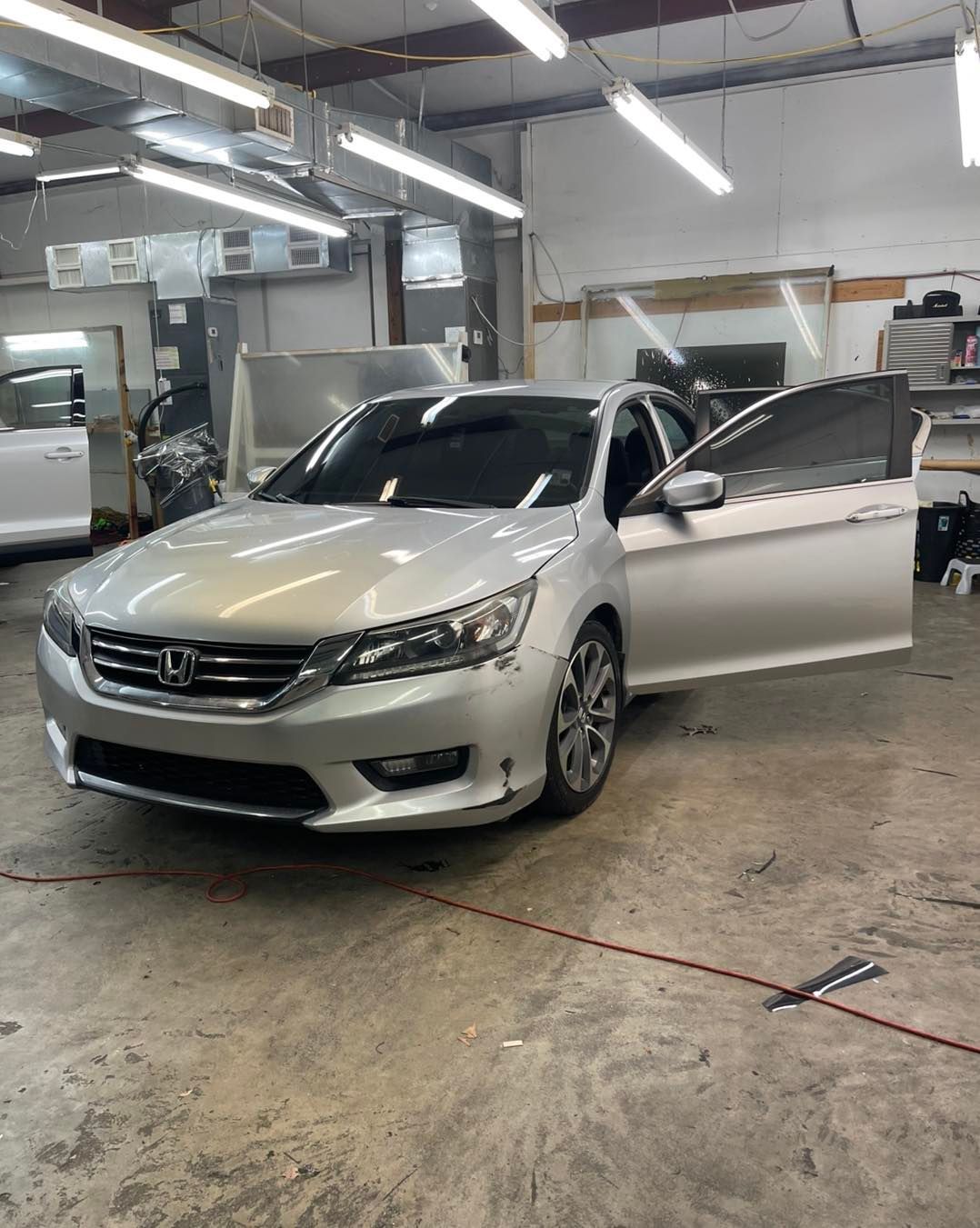 A silver Honda sedan with its driver-side door open inside a brightly lit automotive workshop.