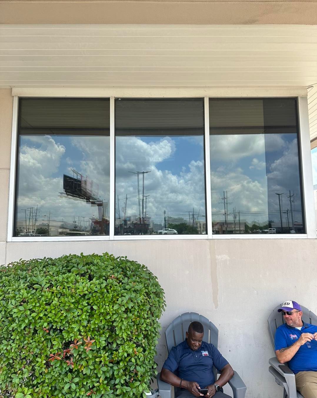 Two people sit in chairs outside a building with a large reflective window and a green bush in the foreground.