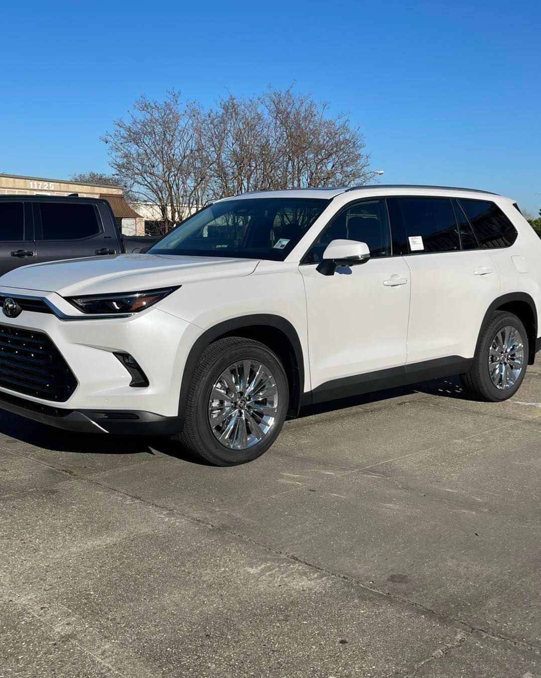 A white Toyota Grand Highlander SUV parked outdoors on a paved lot under a clear blue sky.