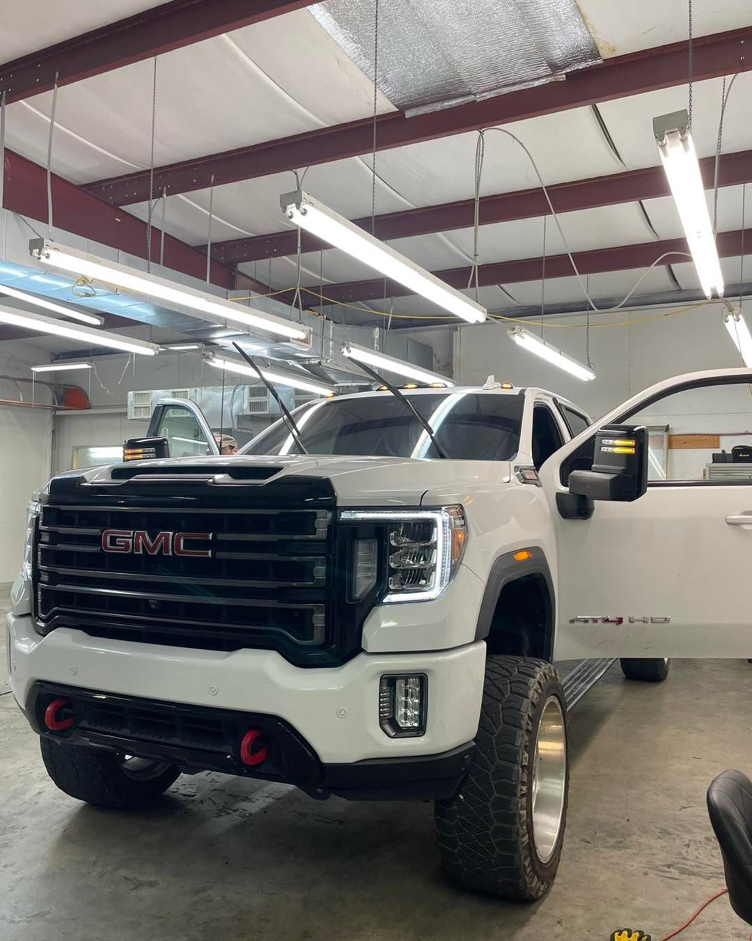 A white GMC pickup truck with a black grille and lifted tires is parked inside a brightly lit automotive workshop.