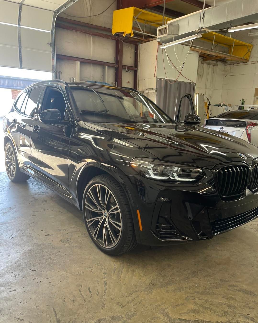 A glossy black BMW SUV parked inside a workshop with concrete floors and industrial overhead equipment.
