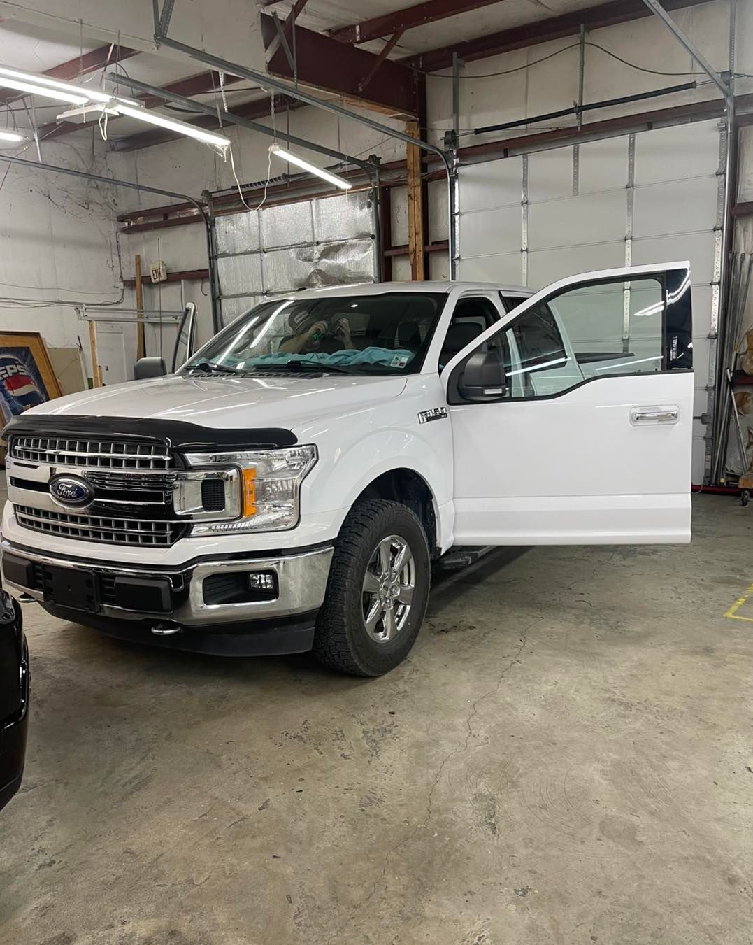 A white Ford F-150 pickup truck with its driver-side door open, parked inside a spacious garage with concrete floors.