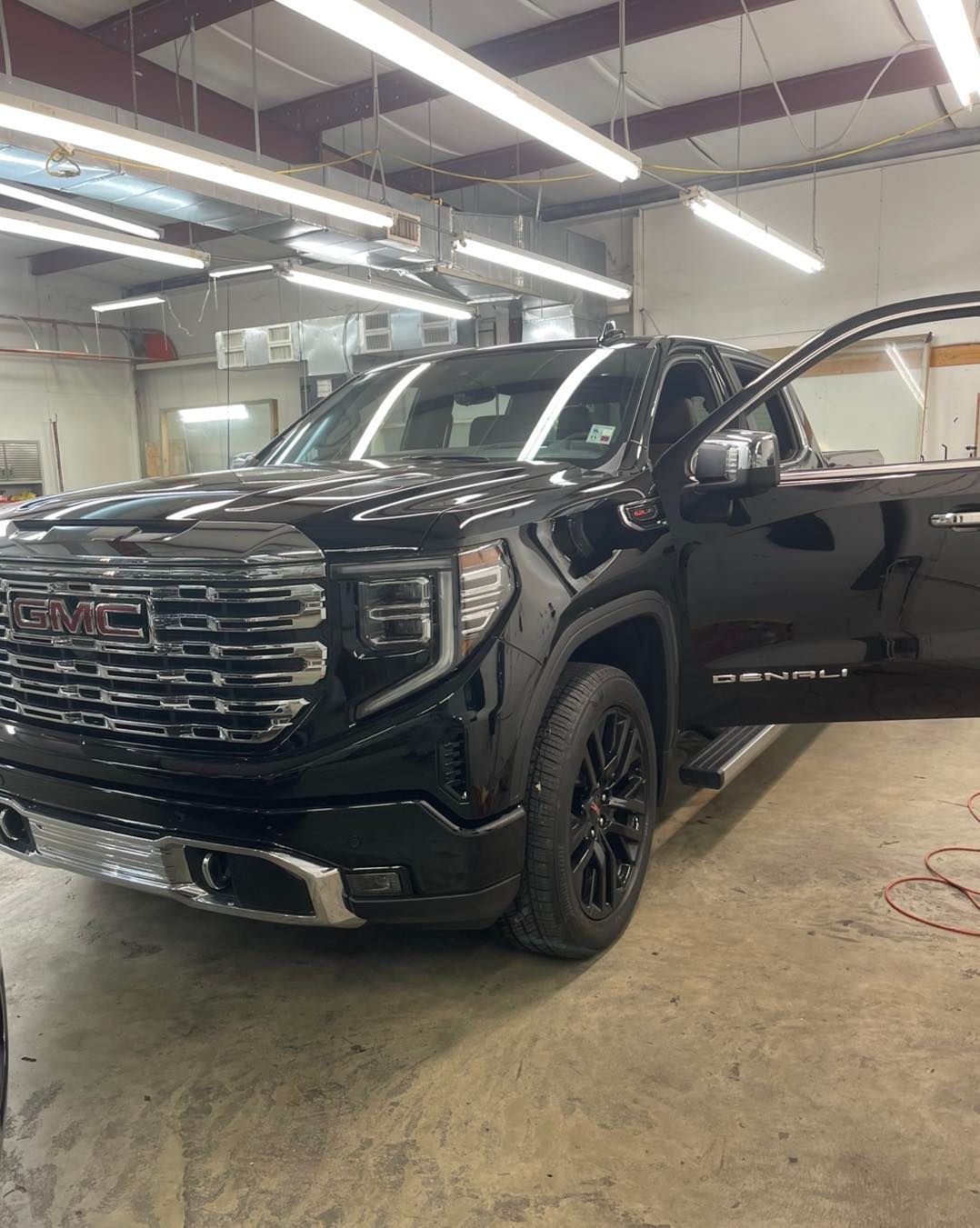 A black GMC Sierra Denali pickup truck parked inside a bright, industrial workshop with its driver-side door open.