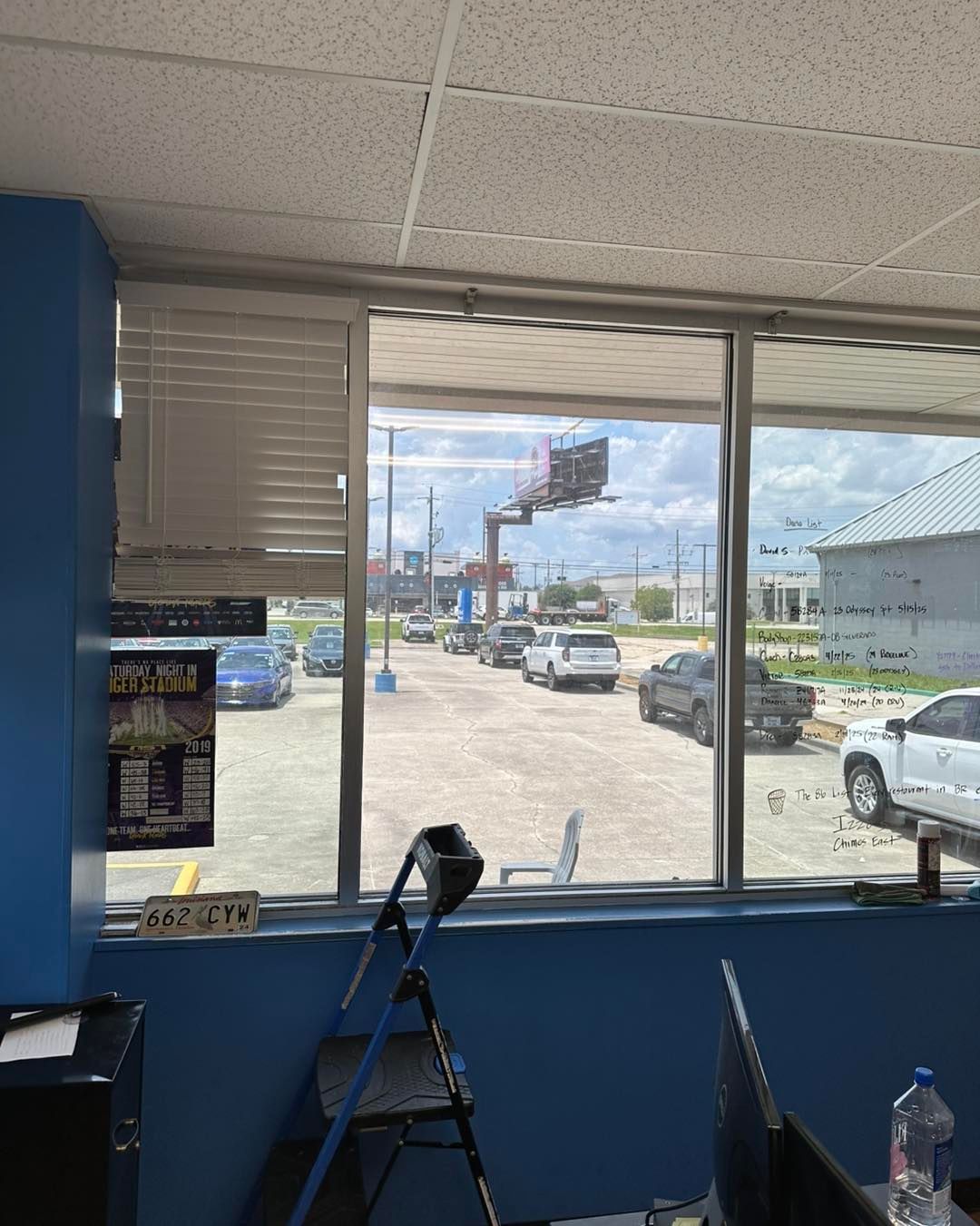 A view from an indoor office window looking out at a sunny parking lot with cars and a billboard under a blue sky.