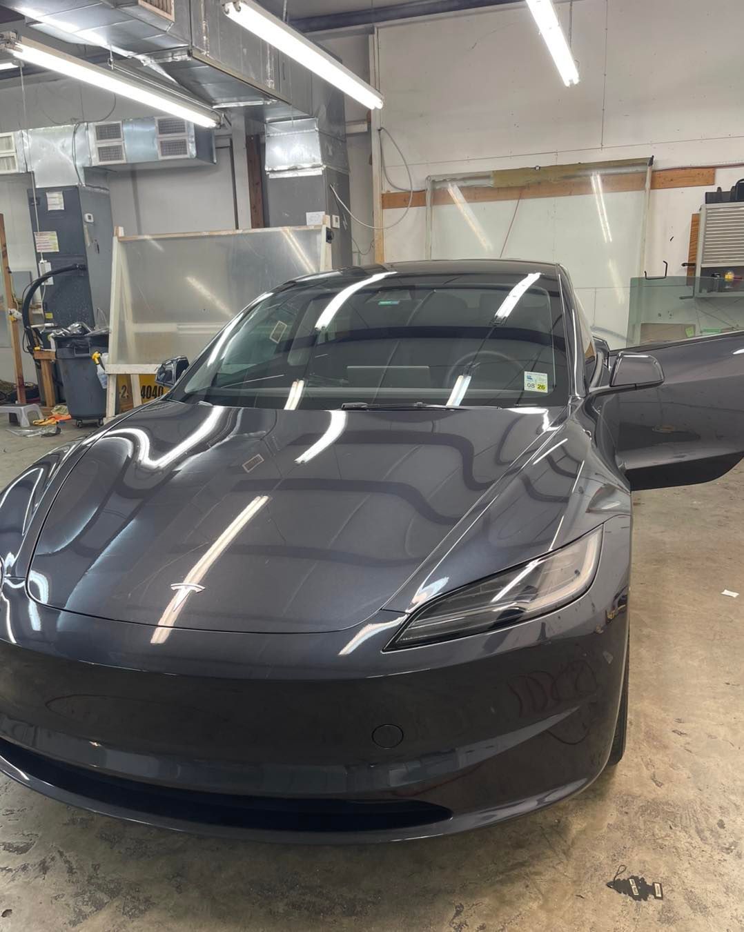 A dark gray Tesla Model 3 parked inside a brightly lit auto shop with the driver-side door open.