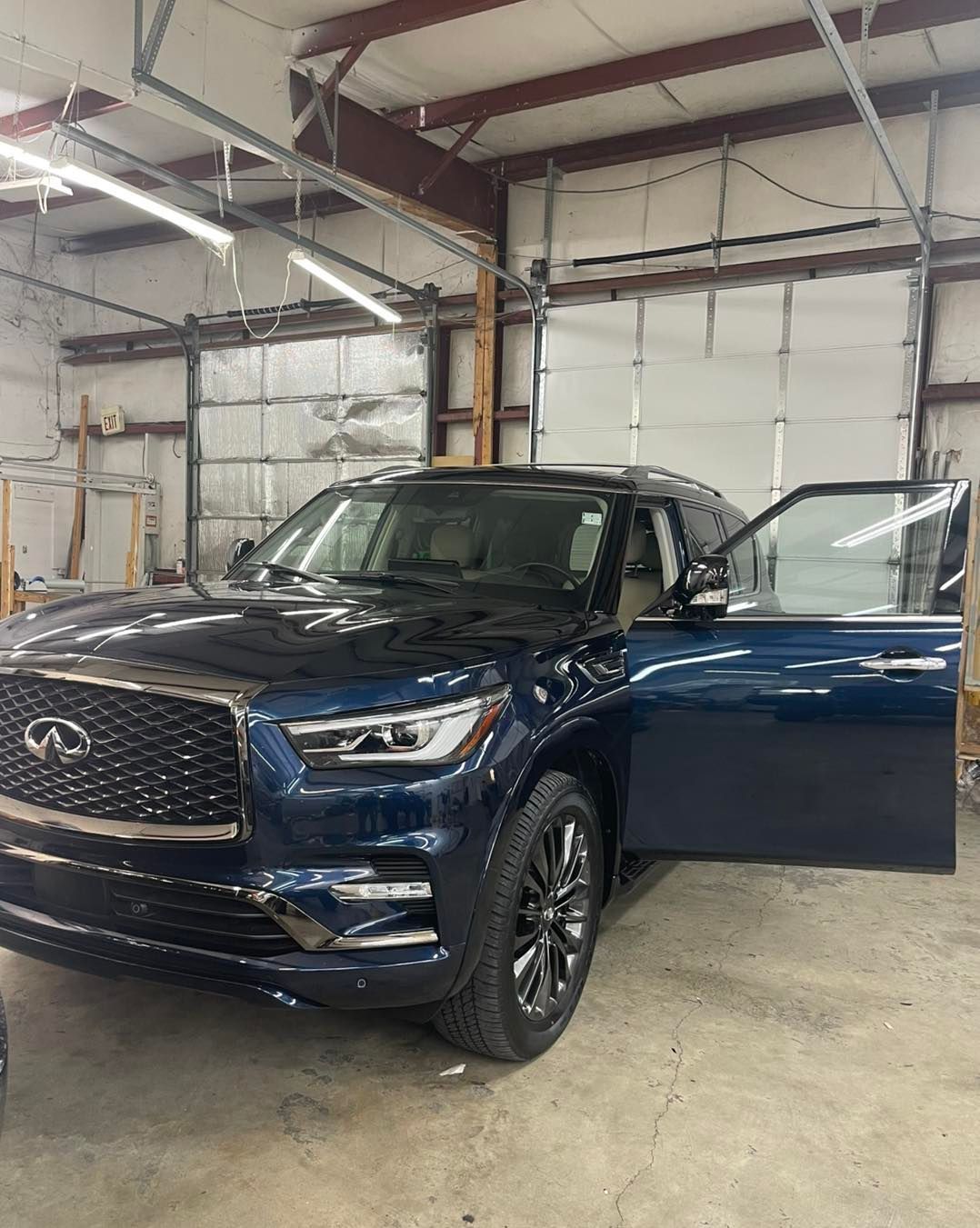 A dark blue Infiniti SUV with its driver-side door open inside a brightly lit garage or workshop.