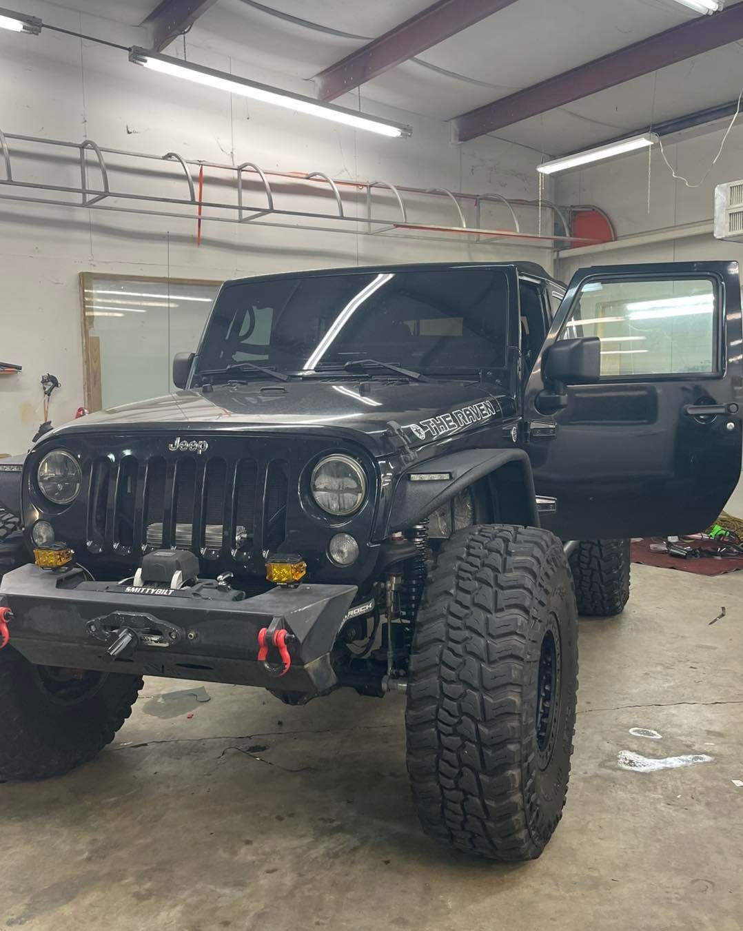Black modified Jeep Wrangler with large off-road tires, winch bumper, and open driver-side door inside a garage.