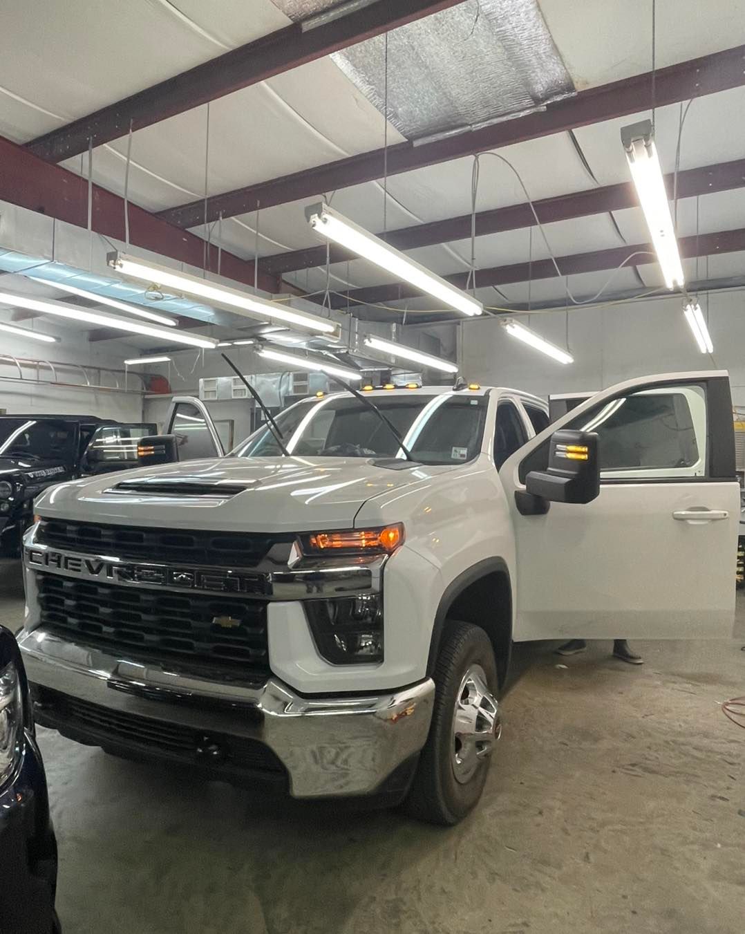 A white Chevrolet dually pickup truck parked inside a bright workshop with its driver-side door open.