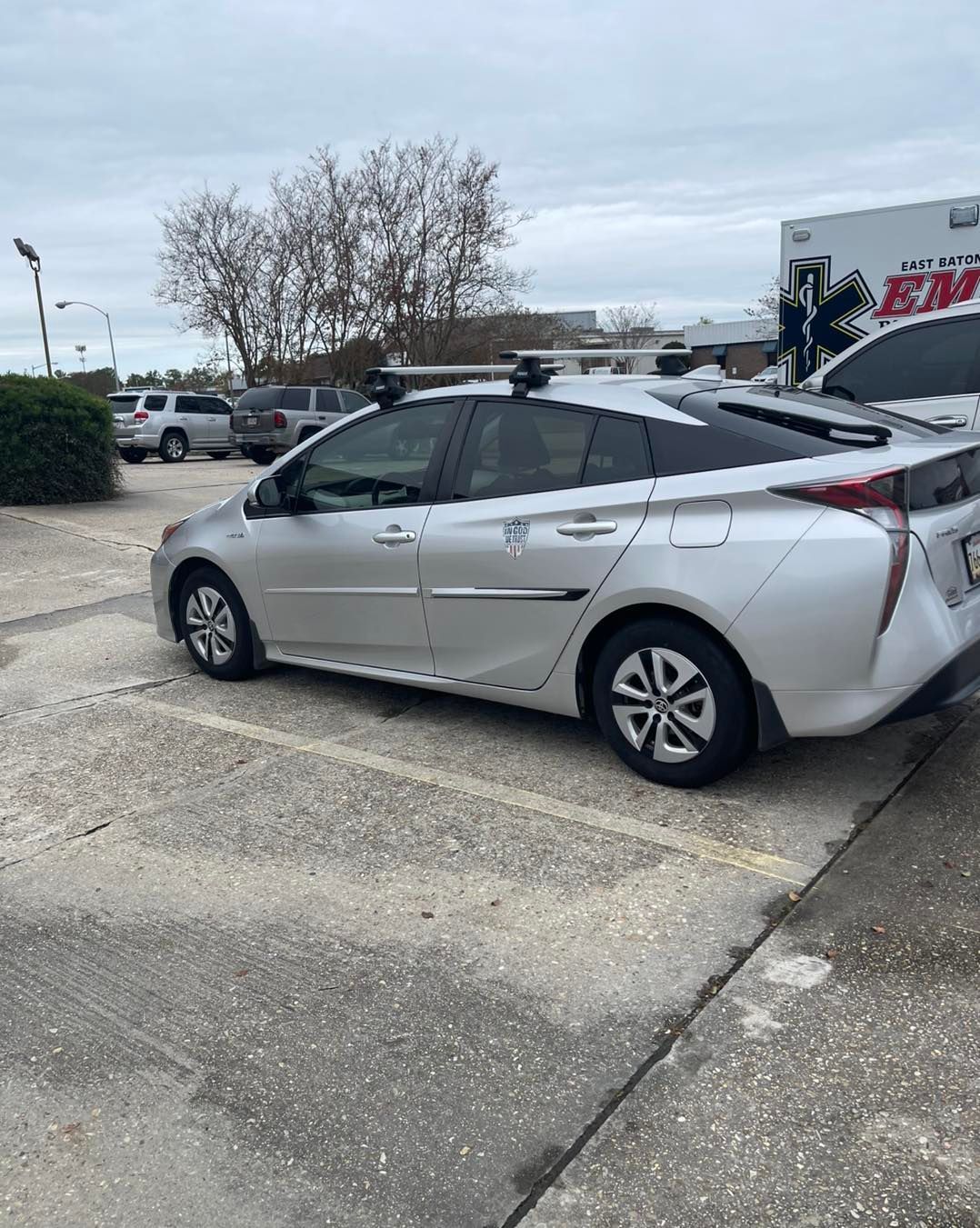 A silver Toyota Prius parked in a paved lot under an overcast sky, with roof racks and a commercial vehicle in the back.