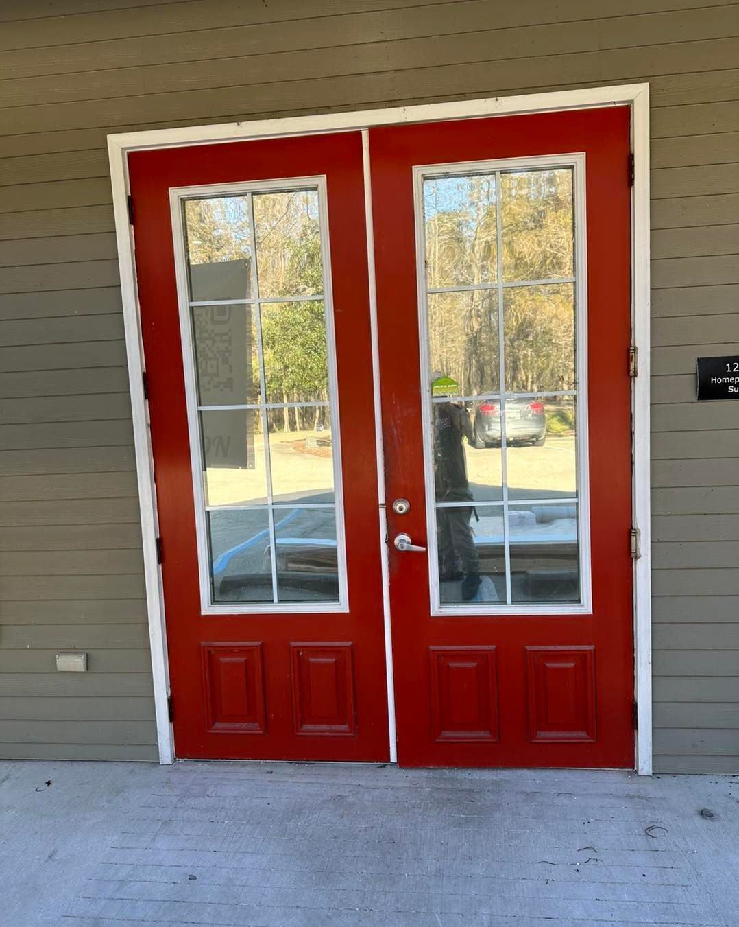 A set of bright red double doors with glass window panes, set against a grey-sided exterior wall on a concrete porch.