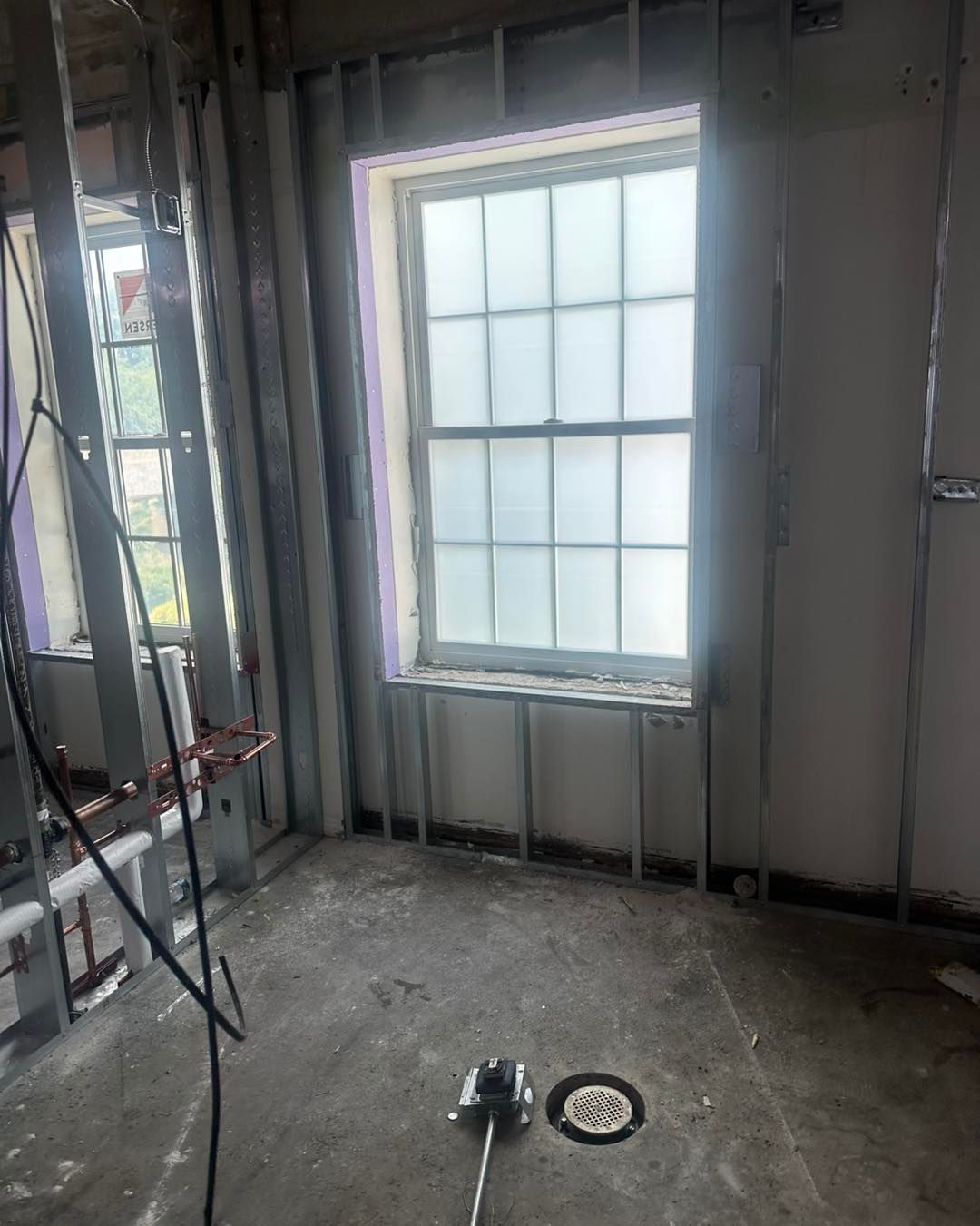 A window in a room under construction, with exposed metal wall studs, concrete flooring, and a floor drain.