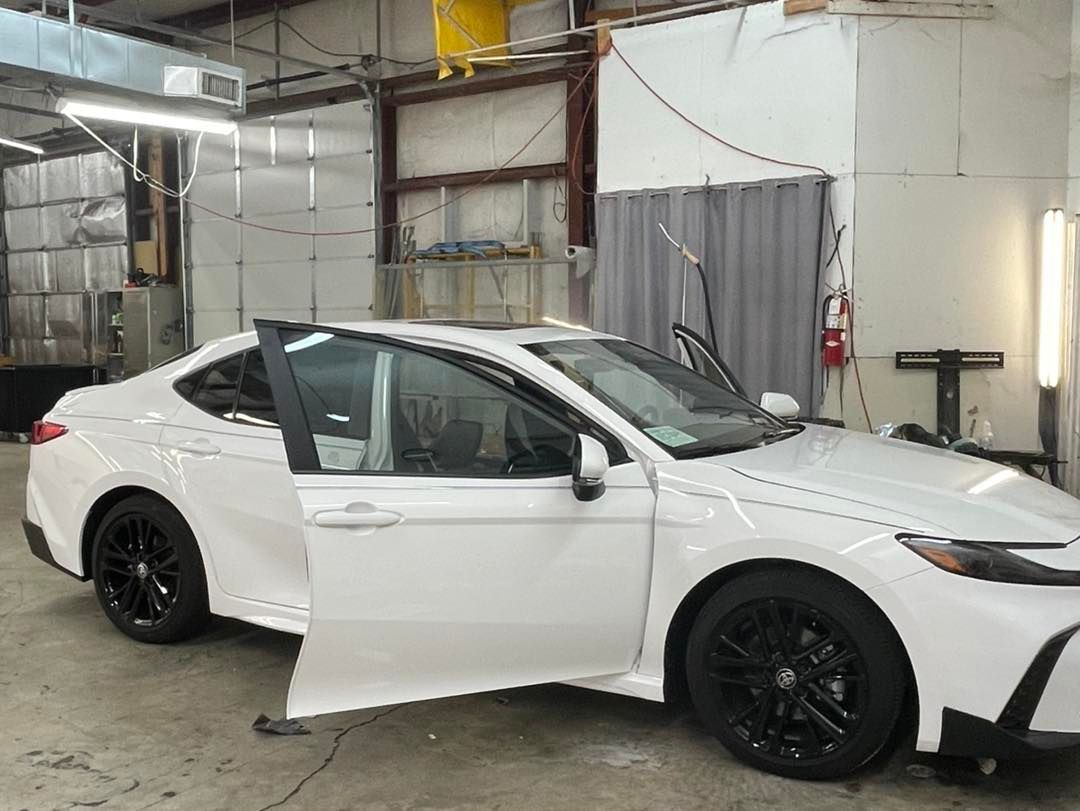 A white Toyota Camry with black rims is parked inside a workshop with its front doors open.