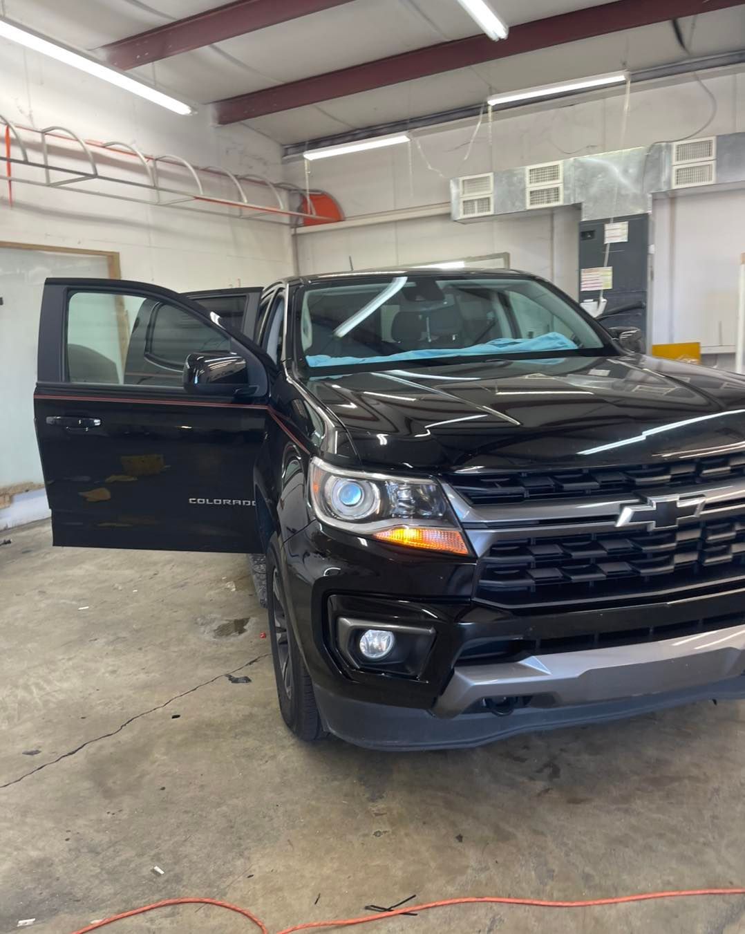 A black Chevrolet Colorado pickup truck parked inside a bright, industrial-style garage with its driver-side door open.