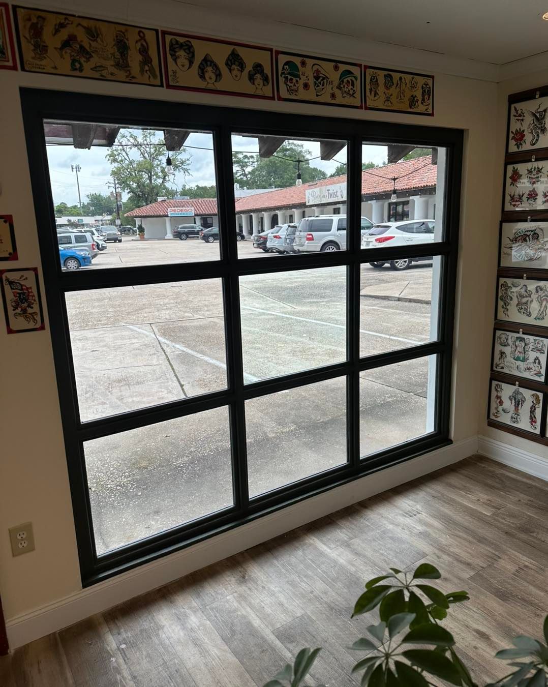 A large black-framed window looks out onto a parking lot with a row of shops, with tattoo art displayed on the walls.