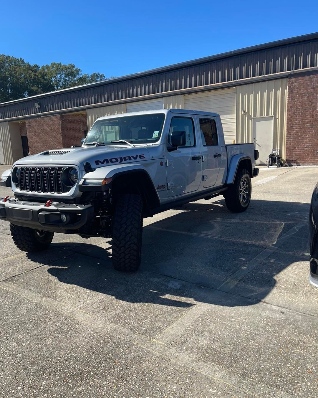 Silver Jeep Gladiator truck with Mojave decals parked on an asphalt lot in front of a brick building.