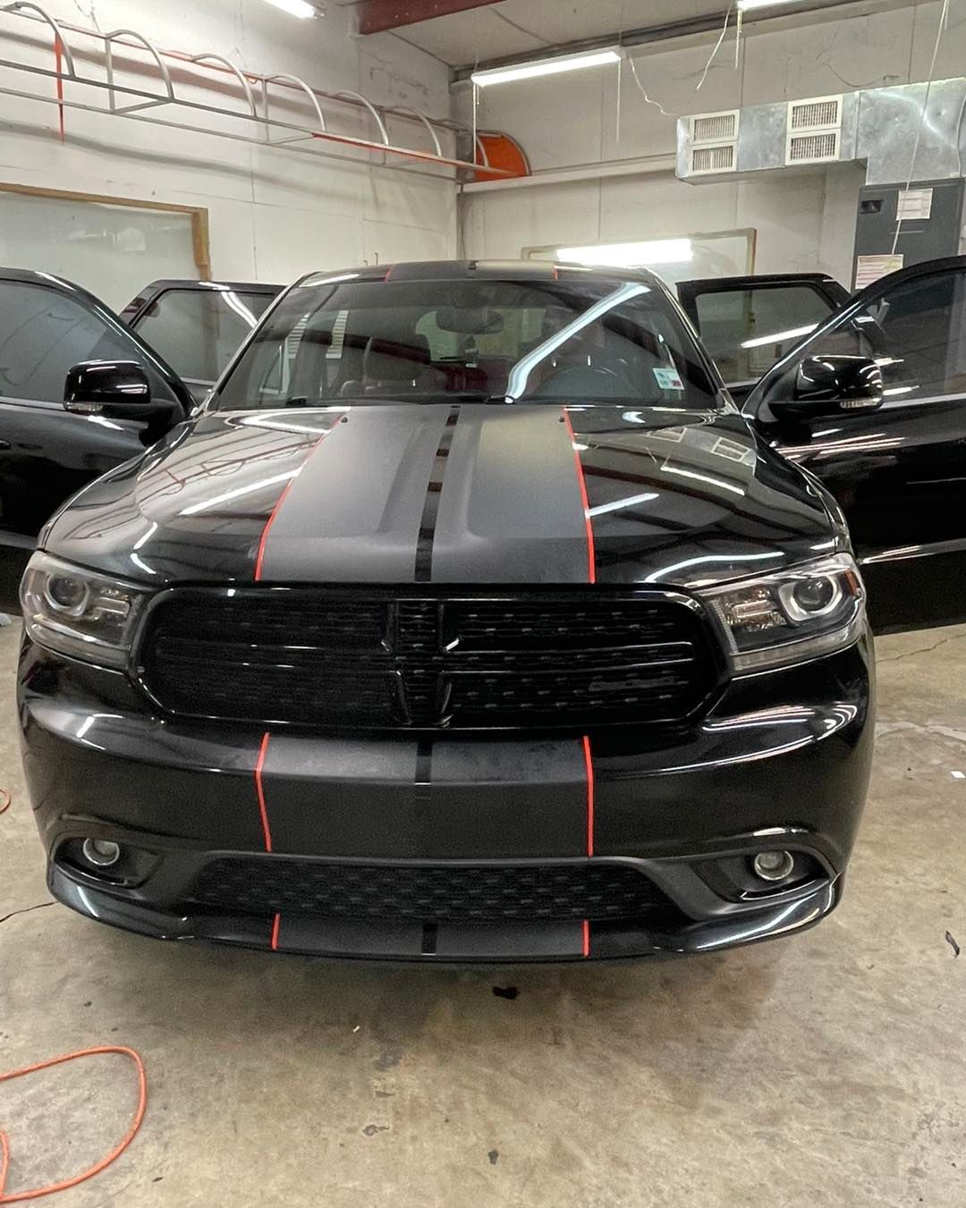 A black Dodge Durango with a matte black and red racing stripe in a garage with open doors.