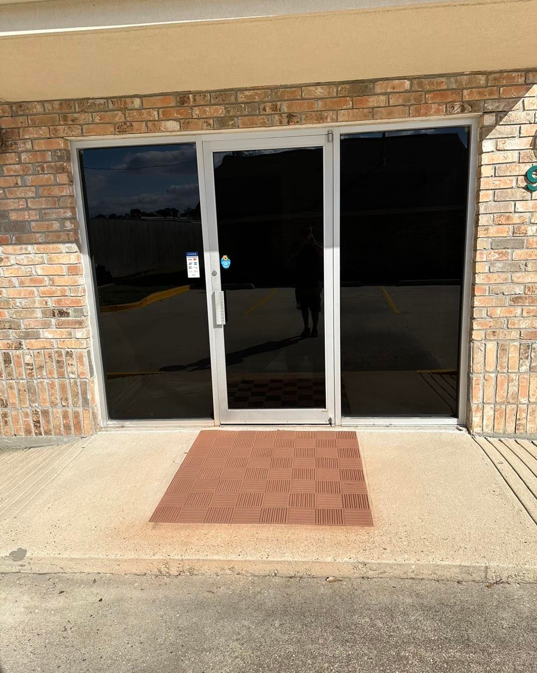 Exterior view of a commercial building entrance featuring a glass door with a brown mat on a concrete walkway.