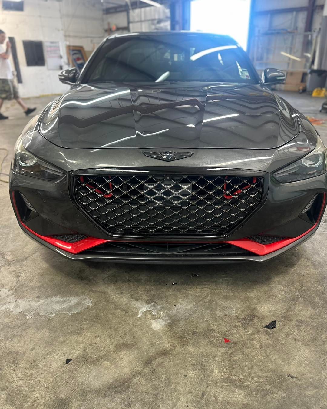 A dark gray Genesis G70 sedan parked in a garage, featuring a black grille and prominent red accents on the front bumper.
