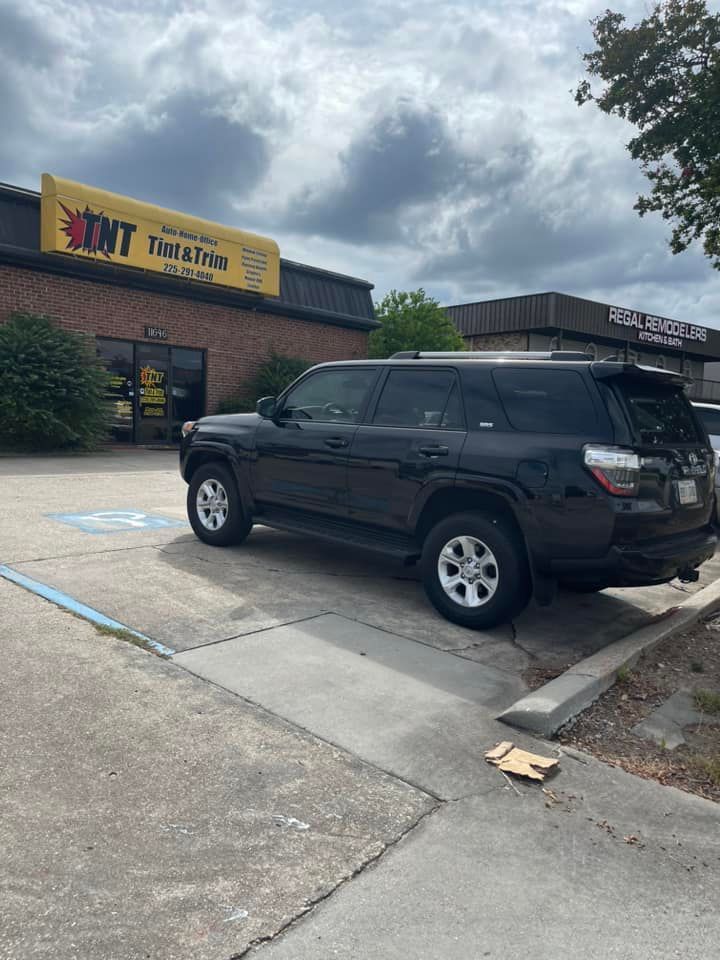 A black SUV parked in an accessible parking space in front of a TNT Tint and Trim shop under a cloudy sky.