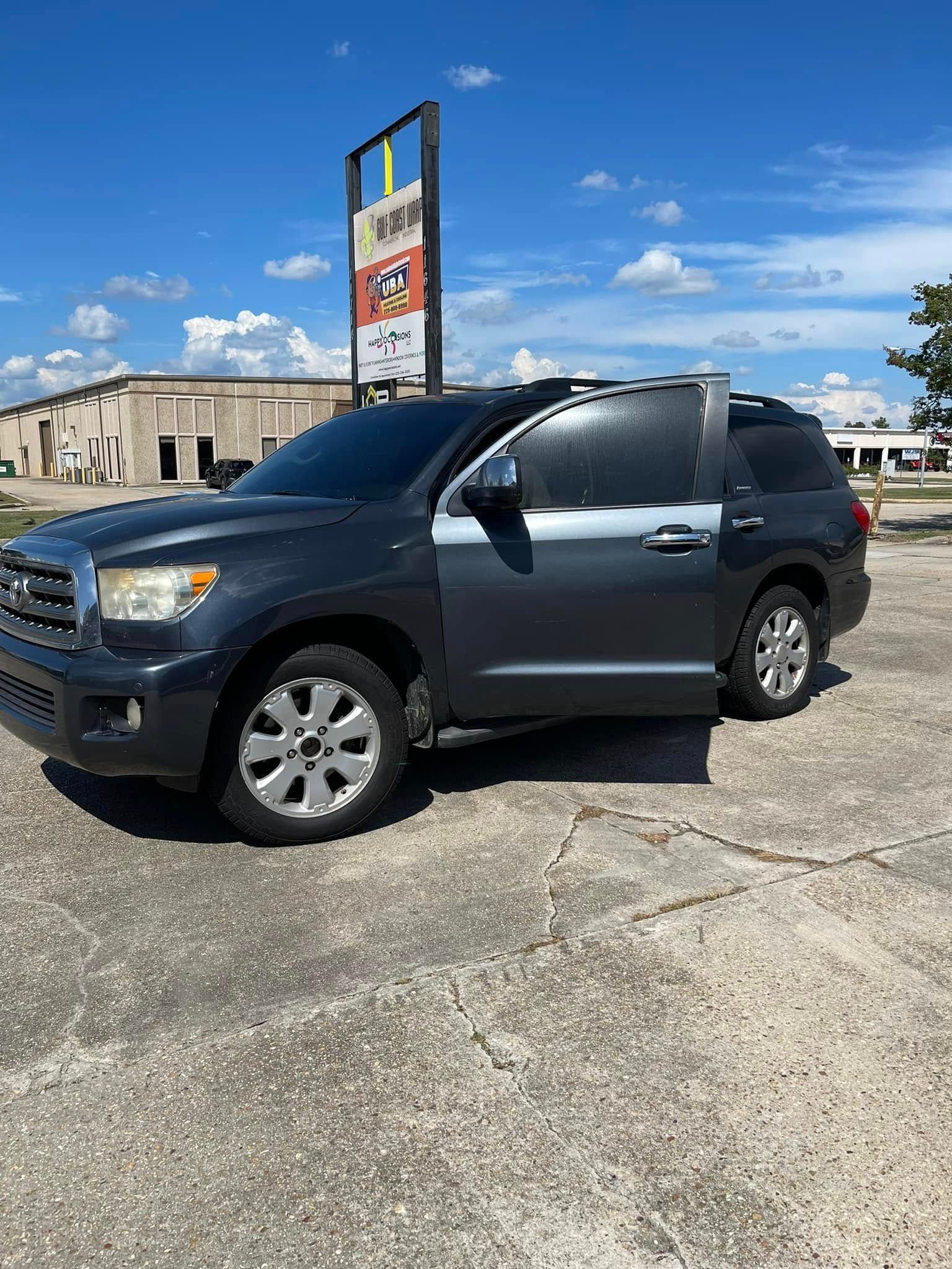 A dark gray Toyota Sequoia parked on a concrete lot under a blue sky, with a tall sign visible in the background.