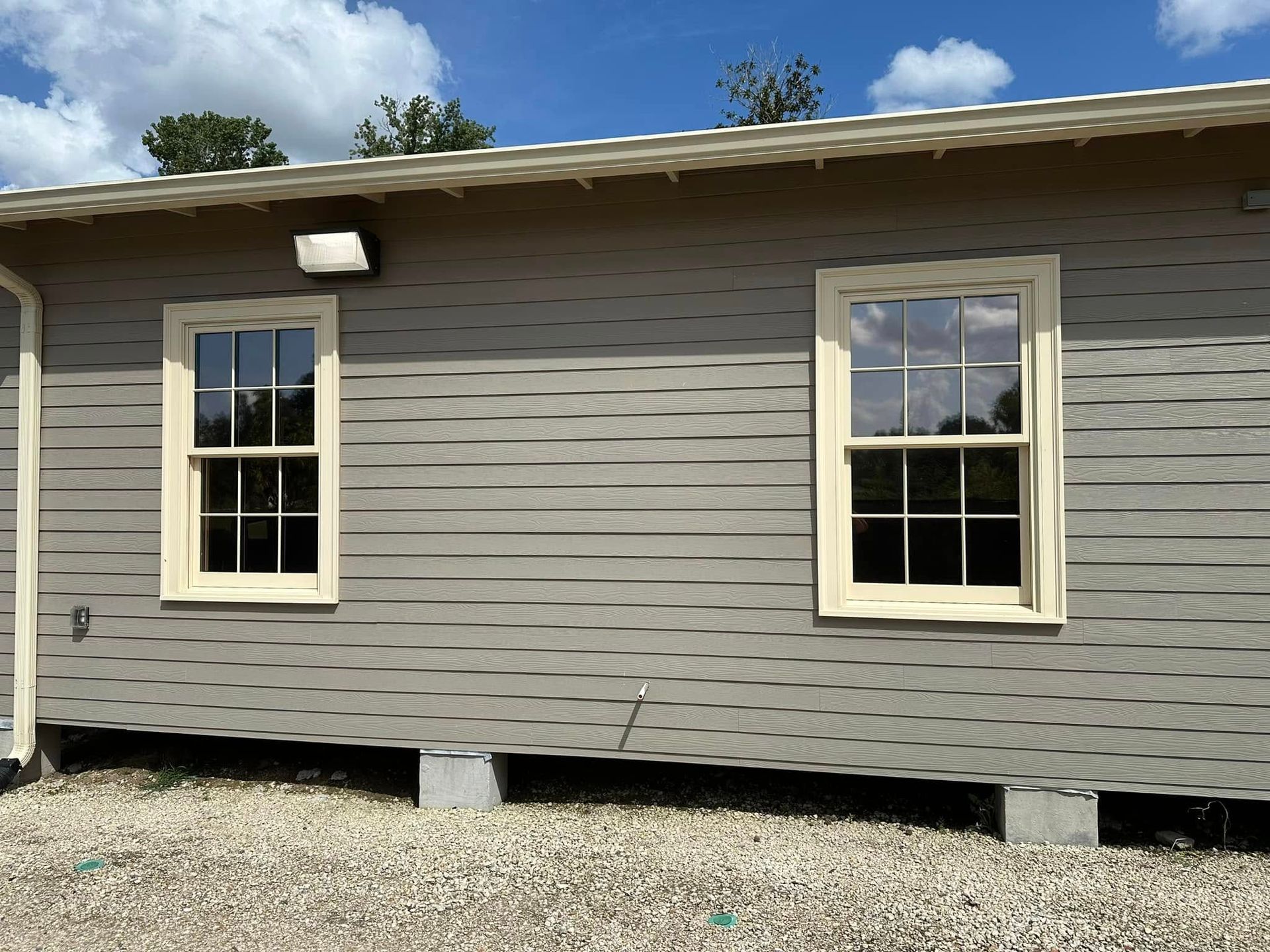 Exterior wall of a gray, horizontal-sided building with two white-framed windows on concrete foundation blocks.