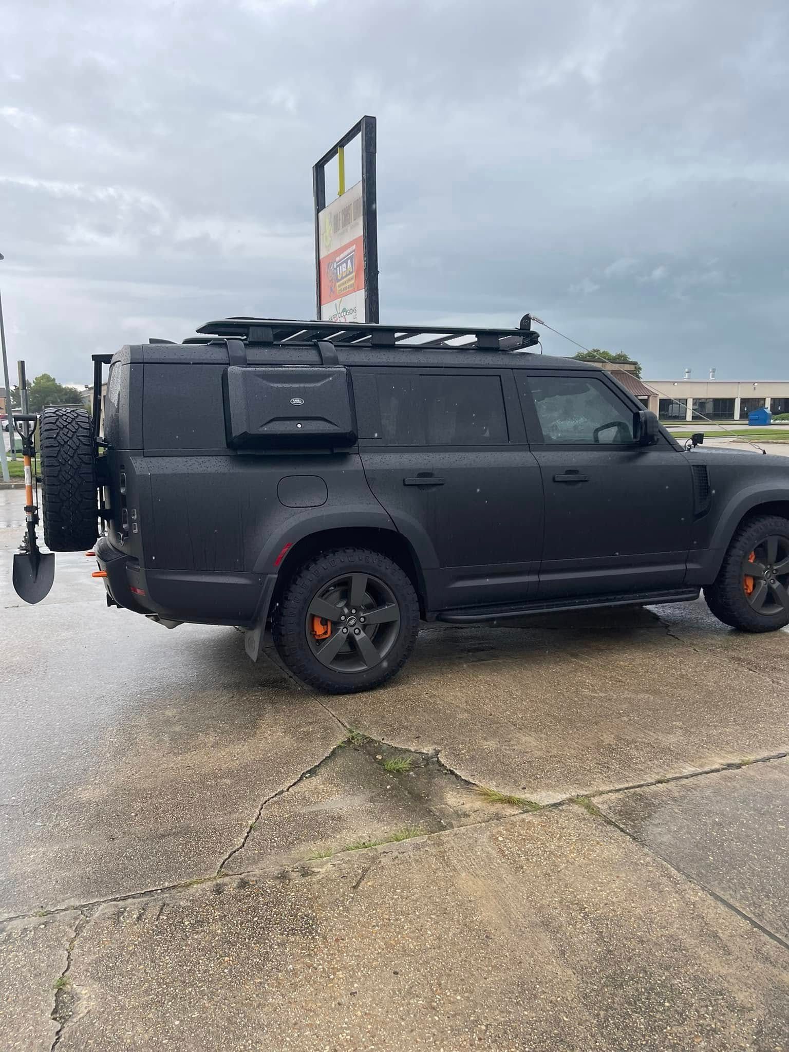 Matte black Land Rover Defender with off-road accessories including a roof rack, storage box, and rear-mounted shovel.