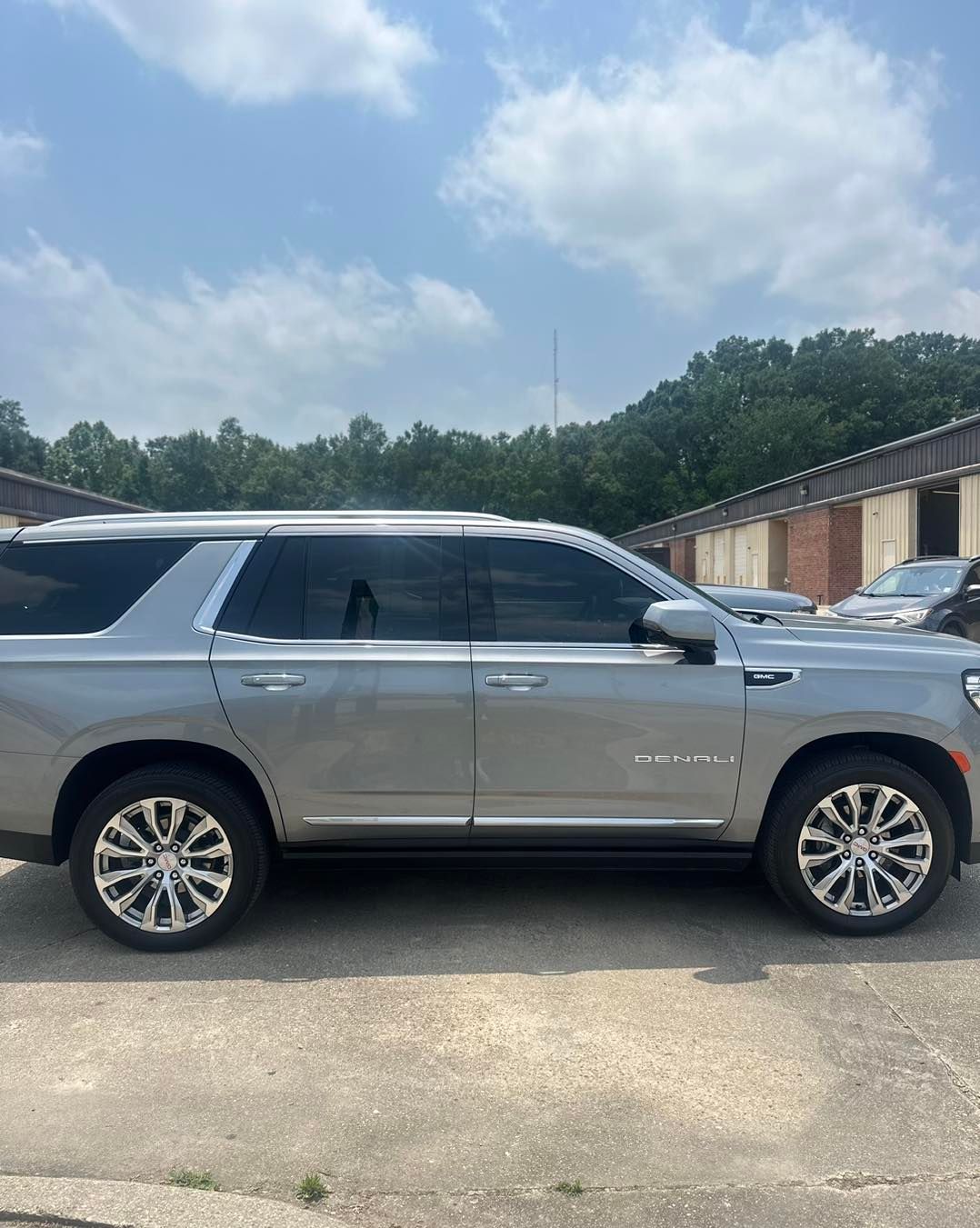 A silver Chevrolet Suburban parked on asphalt in front of a storage facility under a bright, cloudy sky.