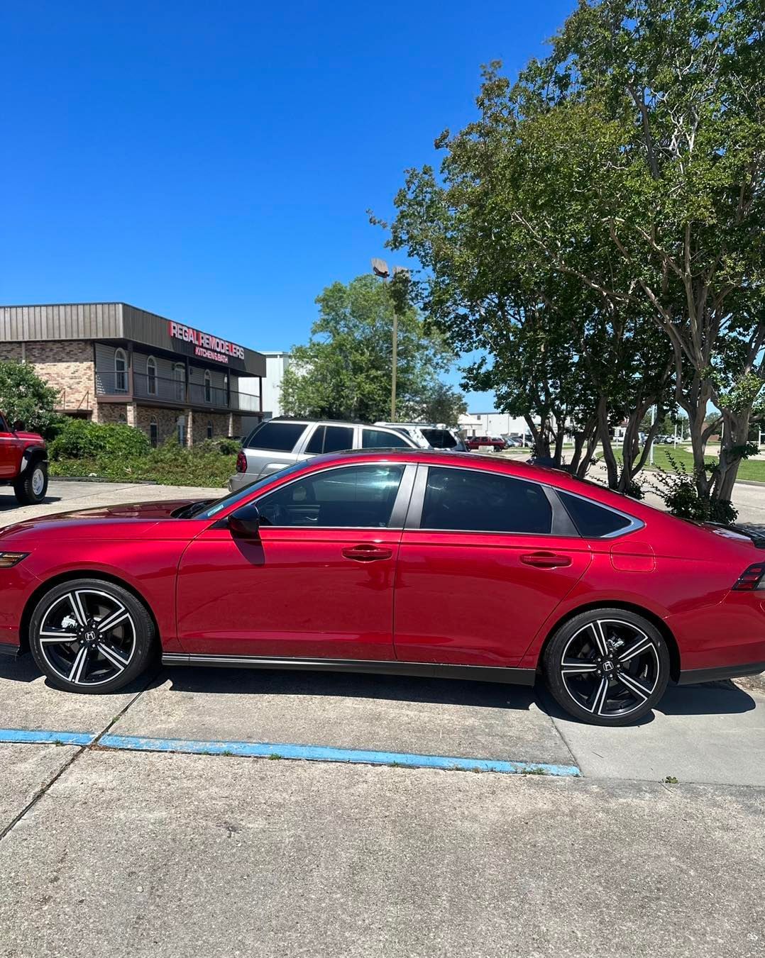 A side view of a shiny red sedan parked in an outdoor lot on a sunny day with a building and trees in the background.