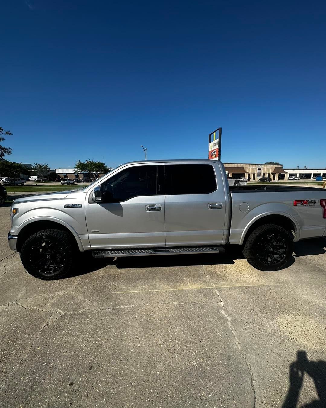 A silver Ford F-150 crew cab pickup truck parked on an asphalt lot under a clear blue sky.